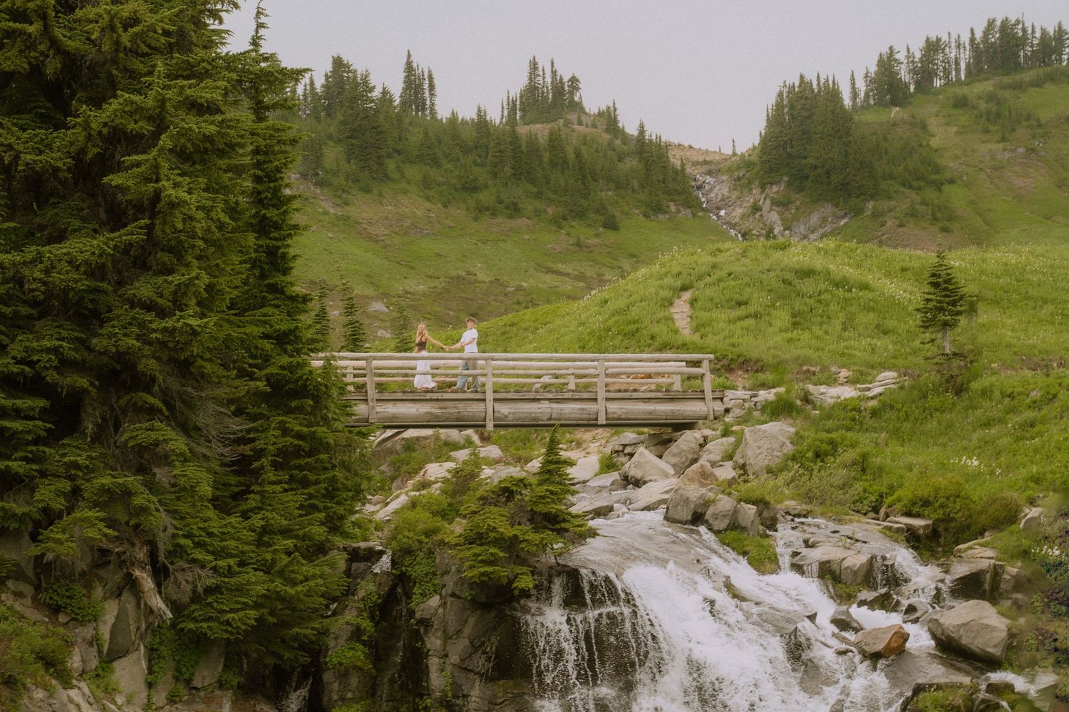 A wooden bridge crosses over cascading waterfalls in a mountainous forest setting.