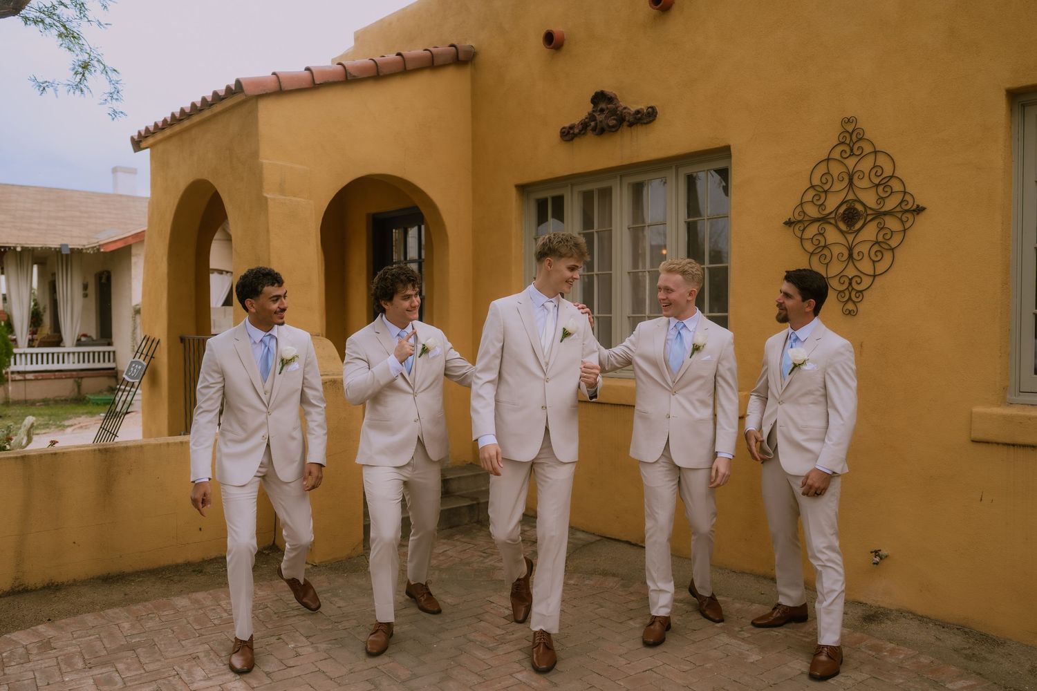 Five people wearing matching white formal attire walk together along a tan stucco wall exterior.