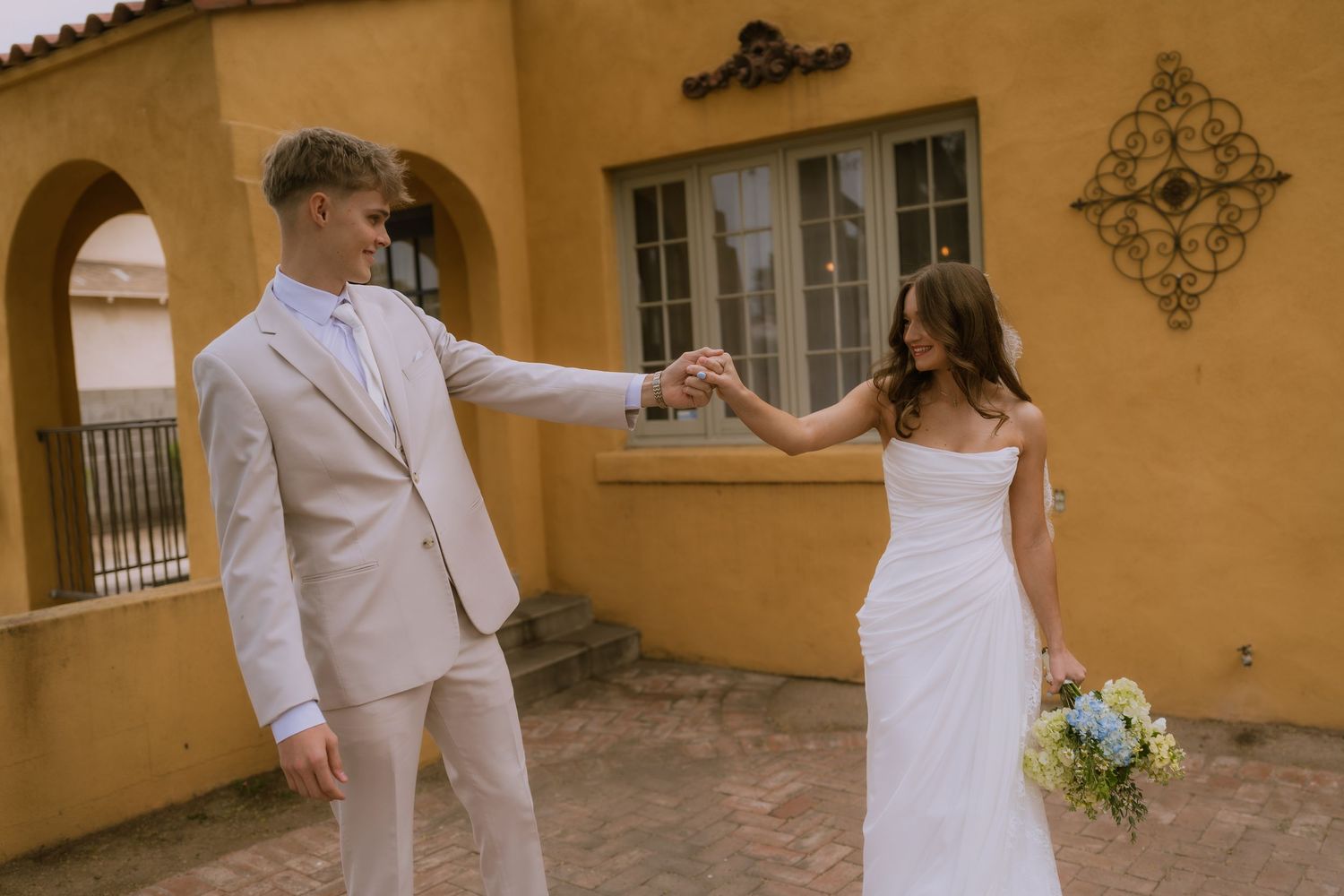 Romantic wedding couple walks along a Mediterranean-style courtyard with peach stucco walls and decorative details.