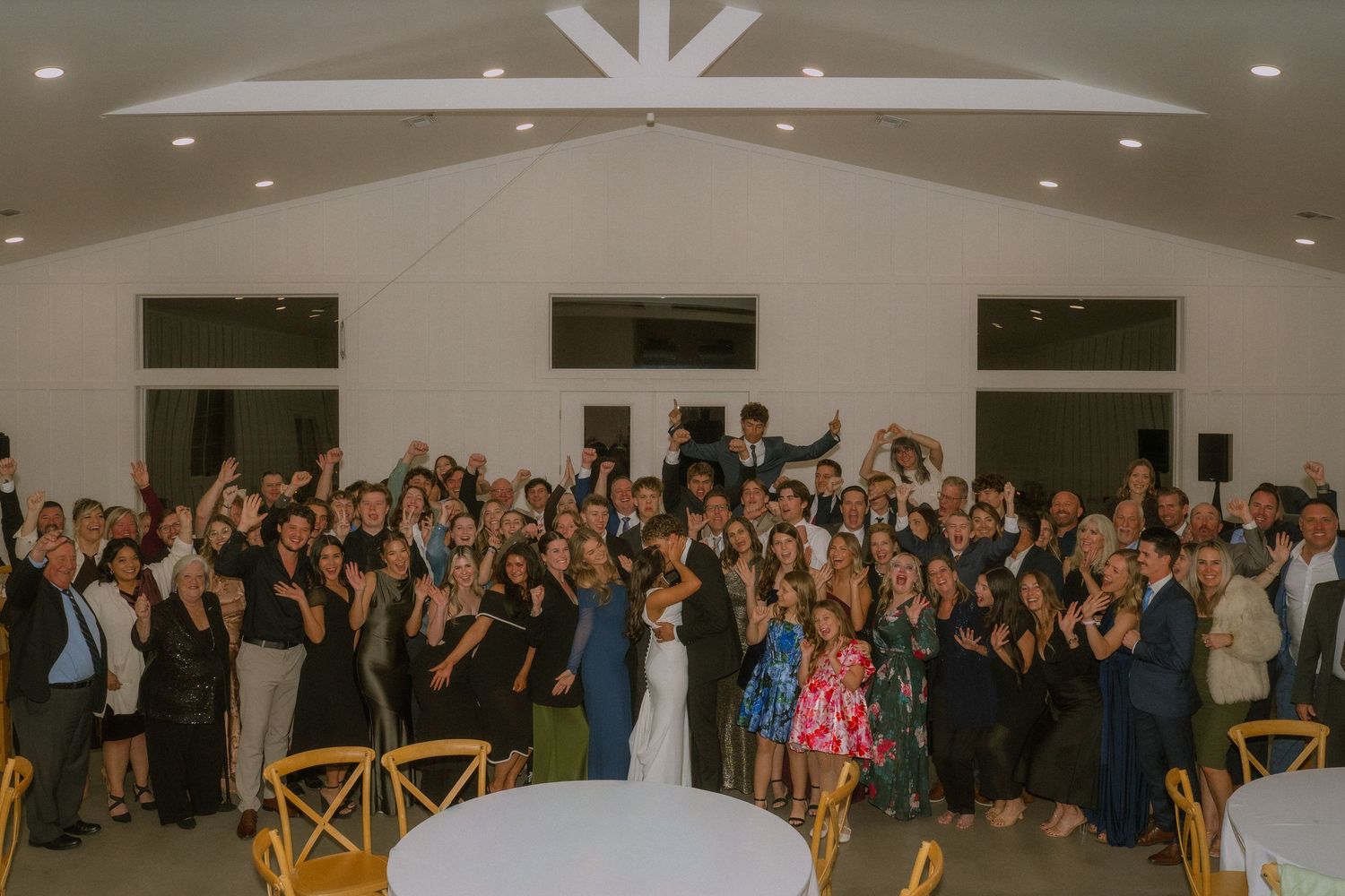 Large group of wedding guests celebrating and dancing together in a brightly lit reception venue.