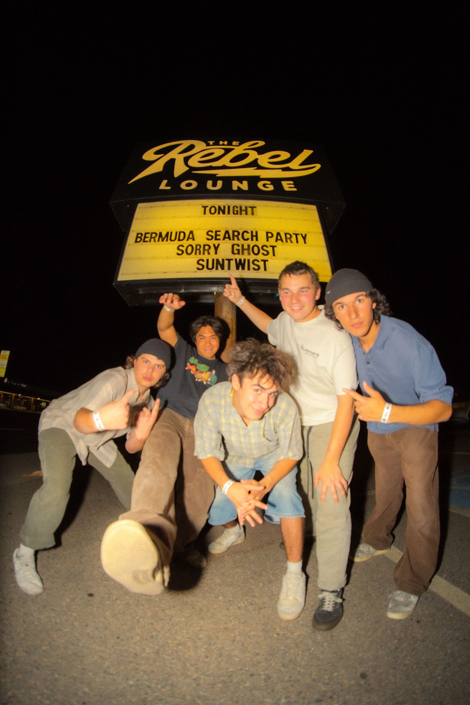 Group poses at night under the Rebel Lounge venue sign advertising a show with Bermuda Search Party and other bands.