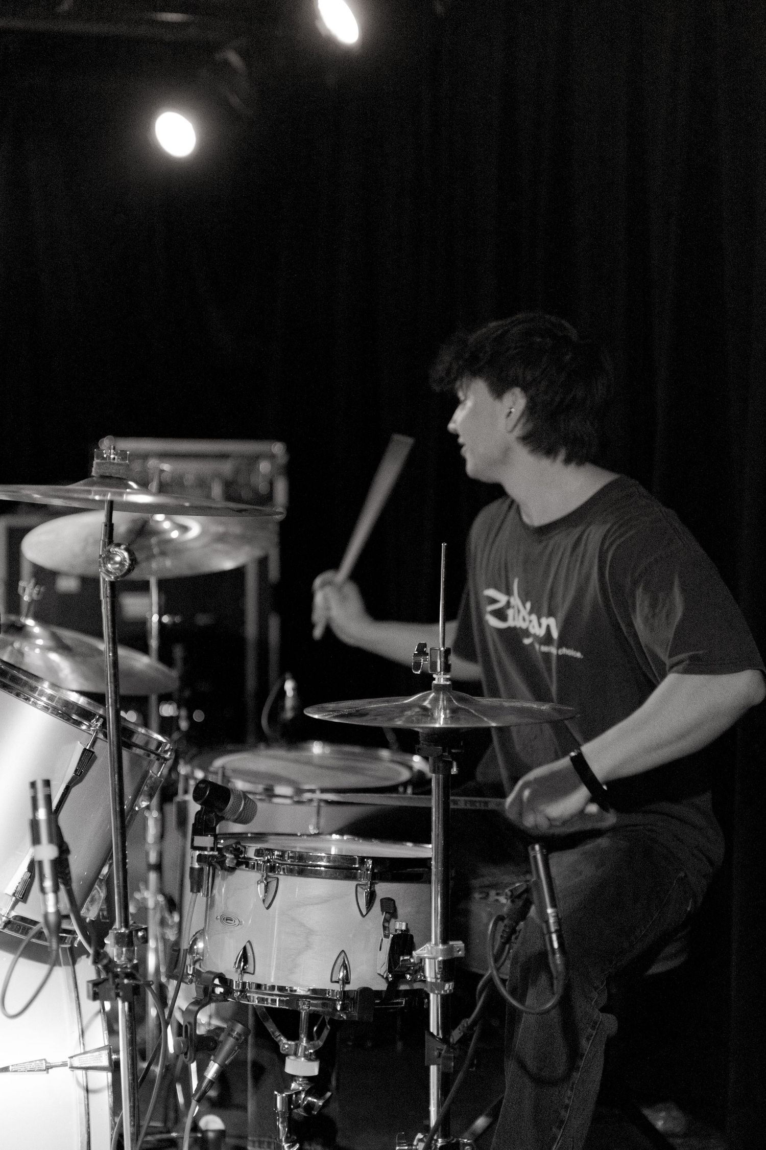A drummer performs on stage in black and white, illuminated by overhead spotlights while playing a drum kit.