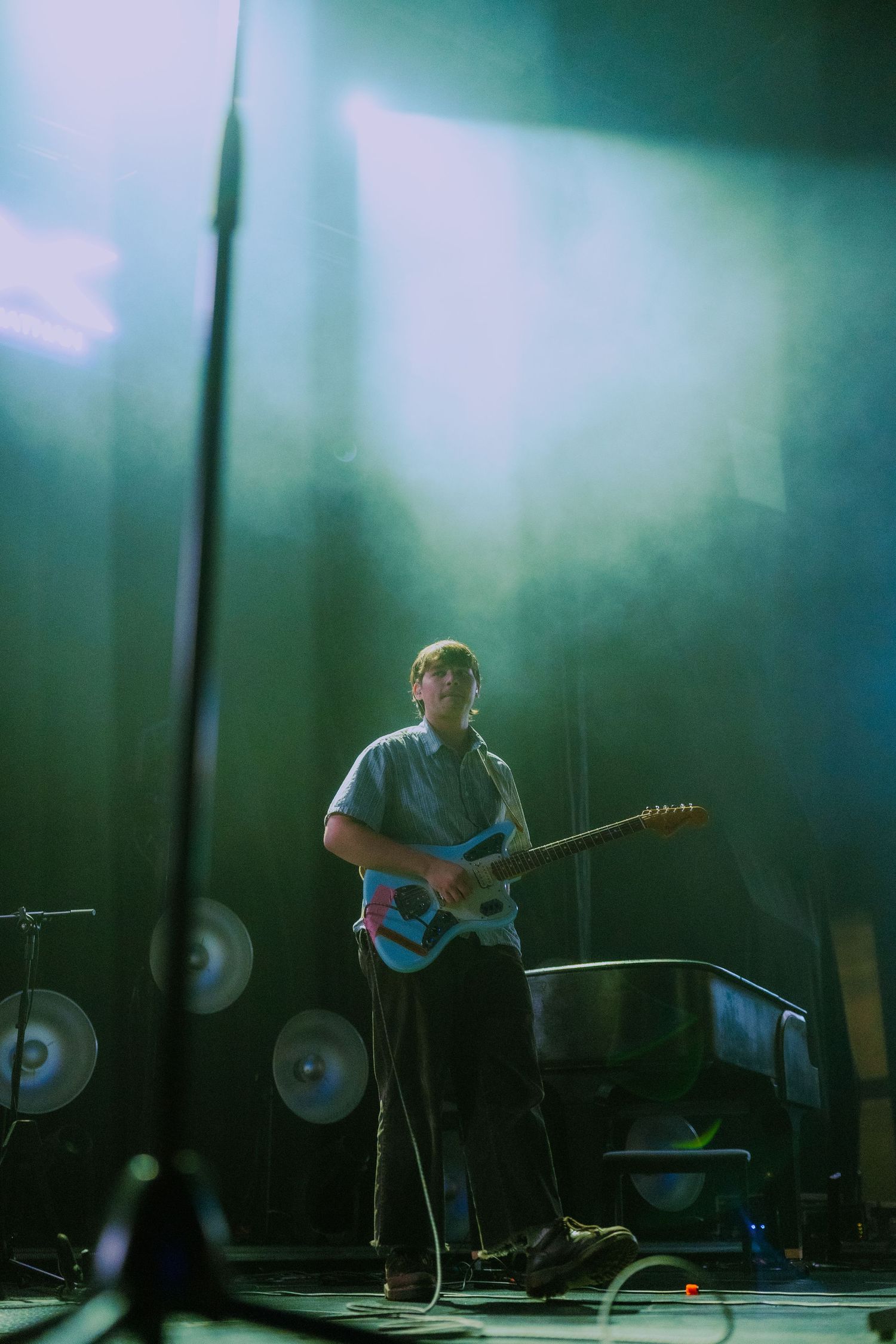 Musician performs on stage with electric guitar in atmospheric lighting with fog and drum kit silhouette in background.