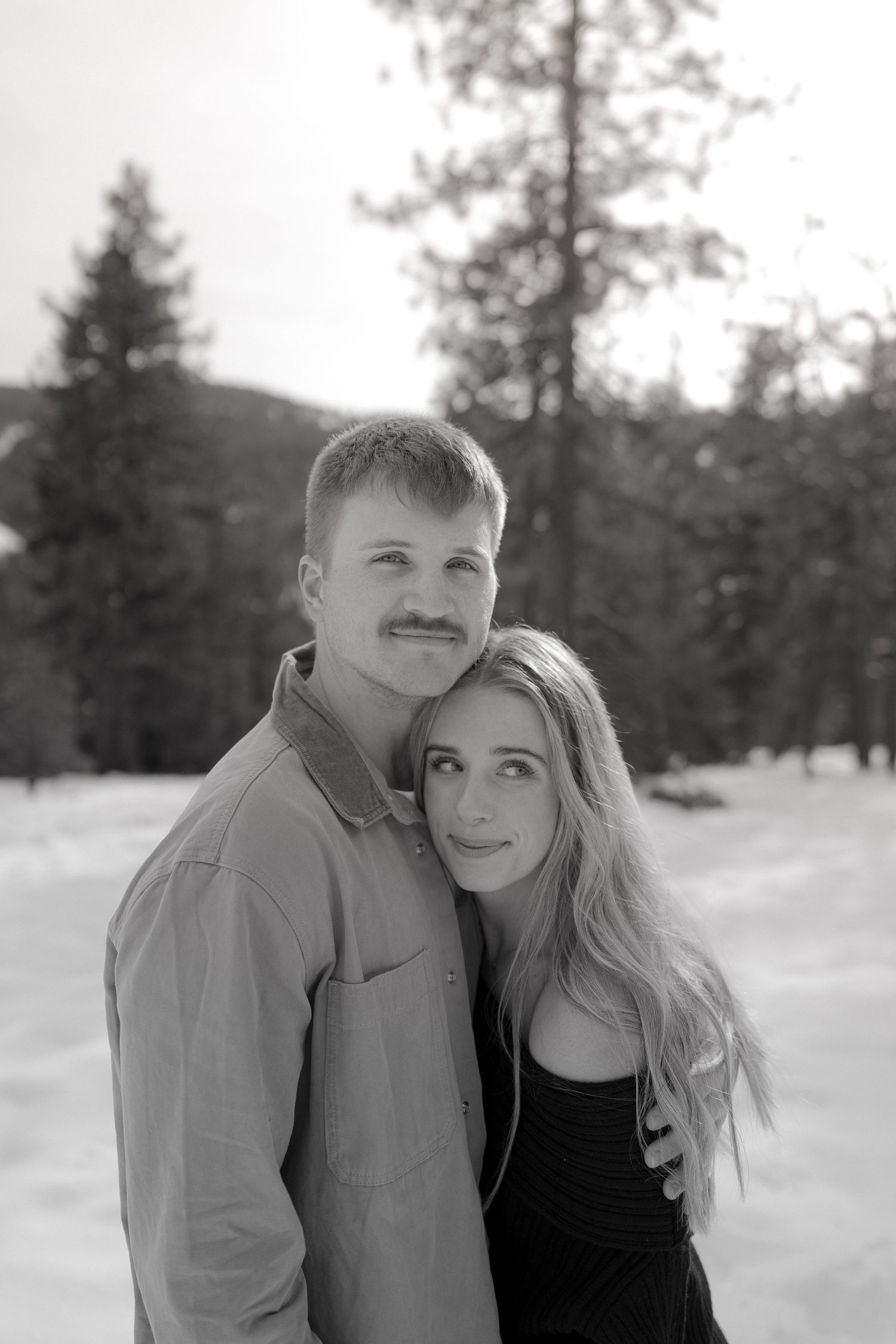 A black and white portrait of a couple embracing outdoors in a snowy forest setting.