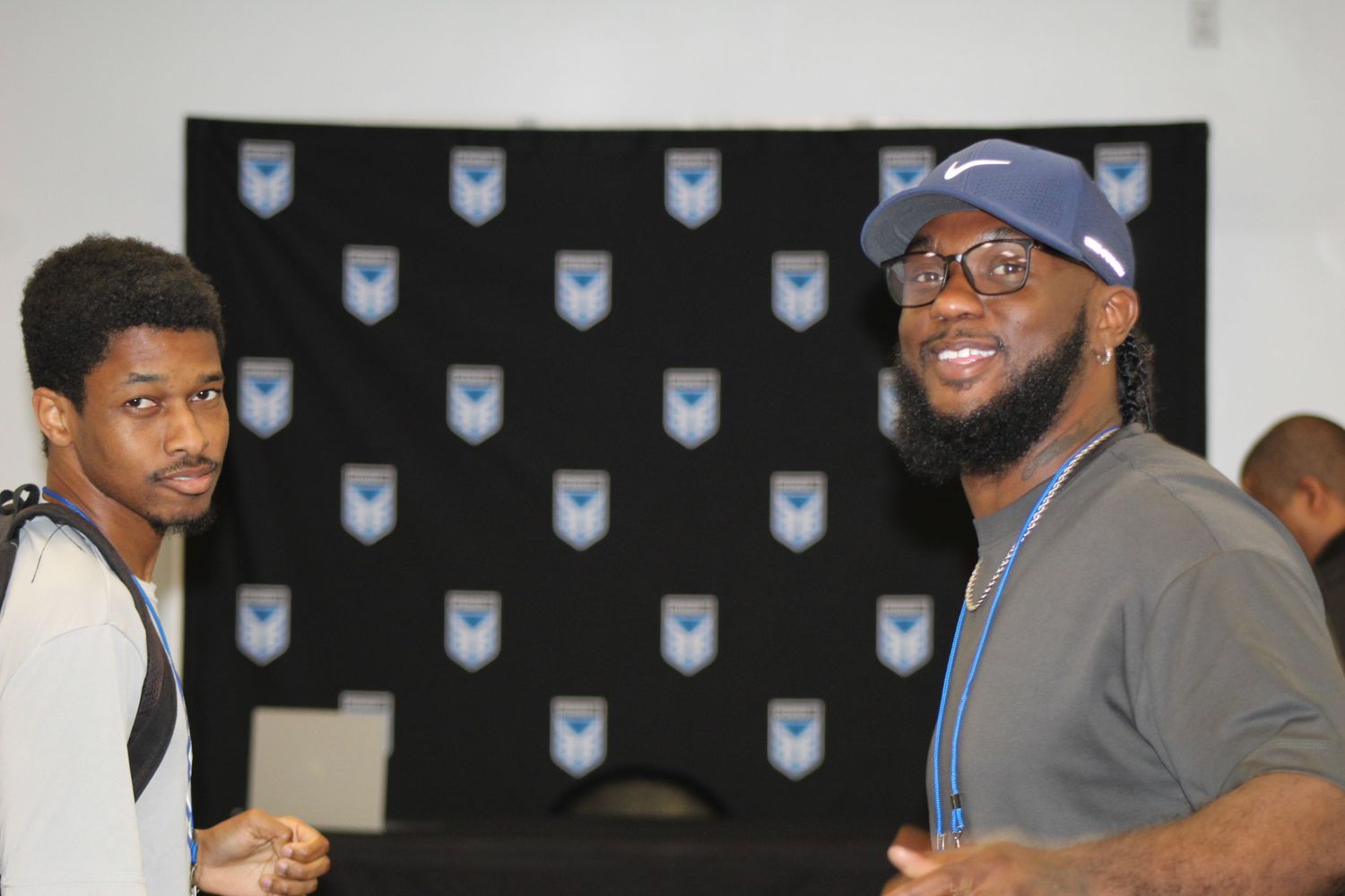 Two men smiling and posing together in front of a black branded backdrop at an indoor event.
