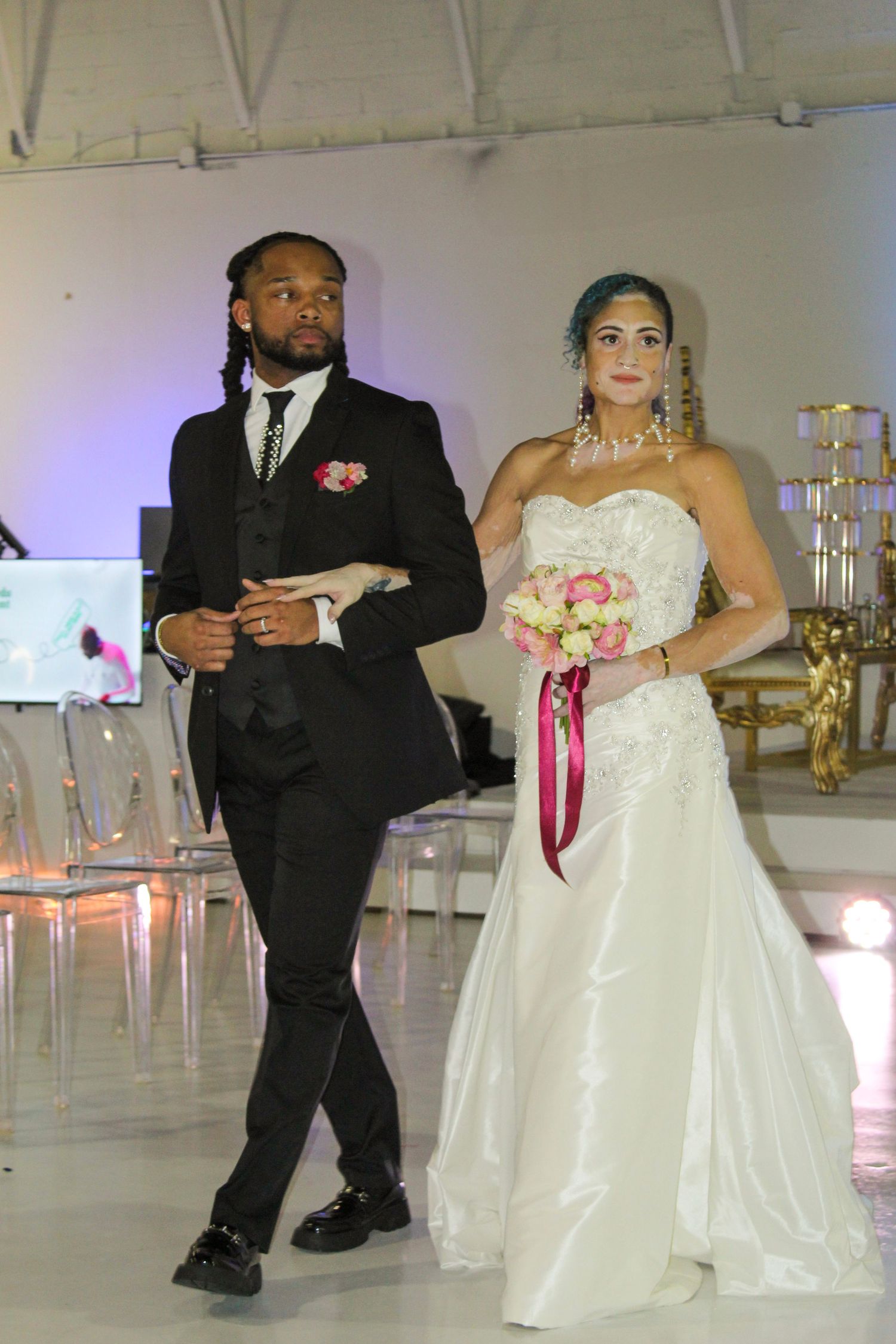 A bride in white gown holding pink bouquet walks with groom in black suit during wedding ceremony entrance.