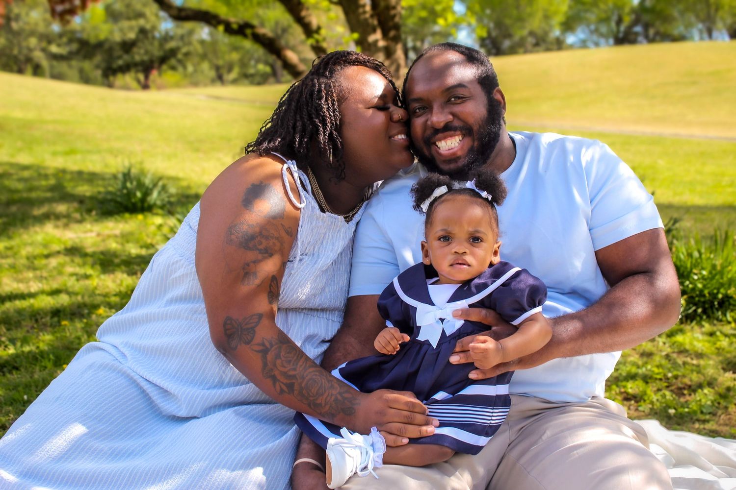 A happy family sits outdoors on grass, with parents smiling and kissing while holding their baby dressed in navy.