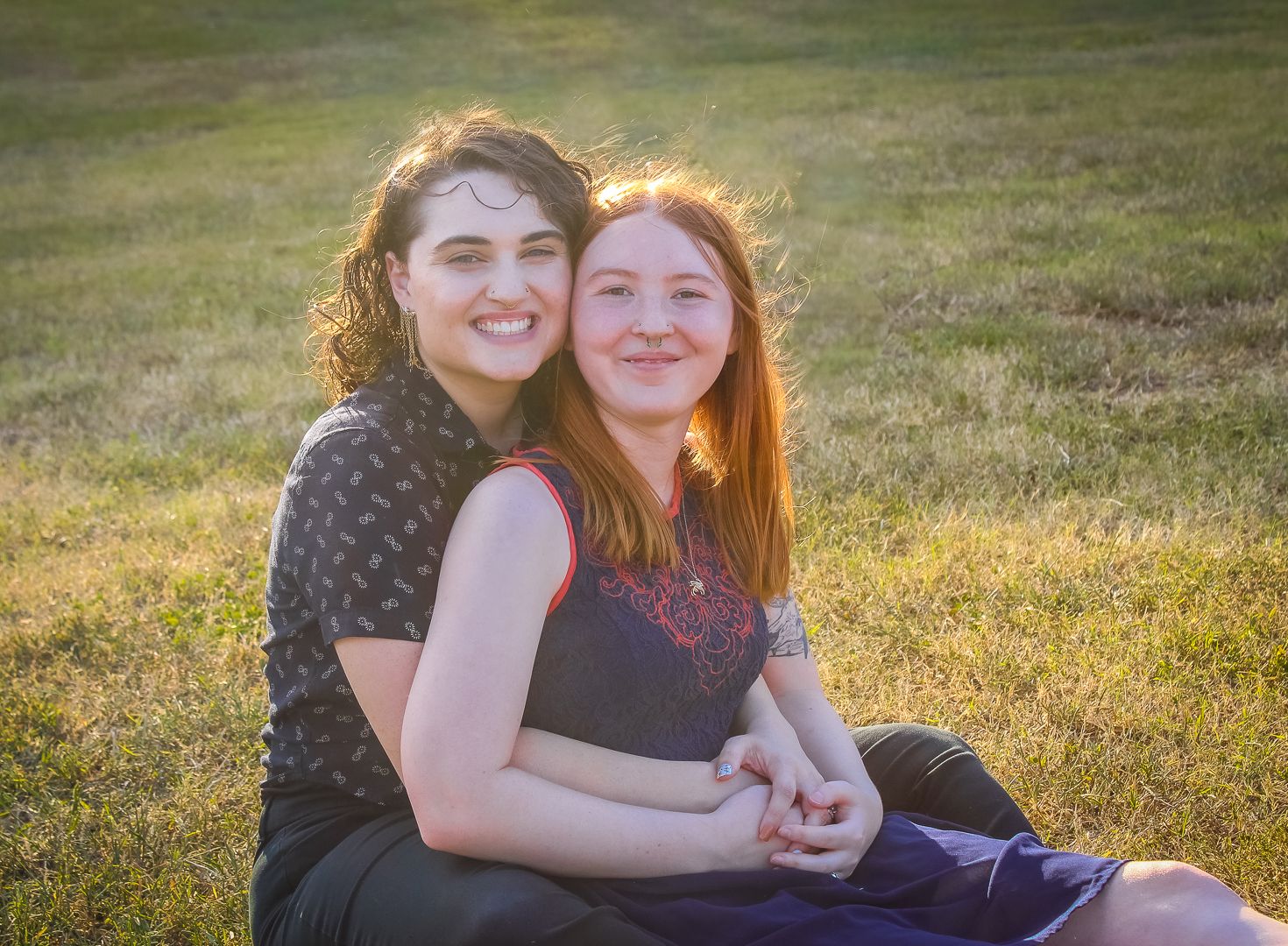 Two young women sitting together on green grass, smiling warmly in golden hour sunlight.