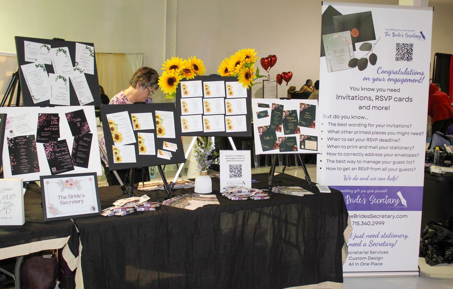 A vendor booth displaying wedding invitations, RSVP cards, and stationery with sunflowers and a promotional banner at a bridal expo.