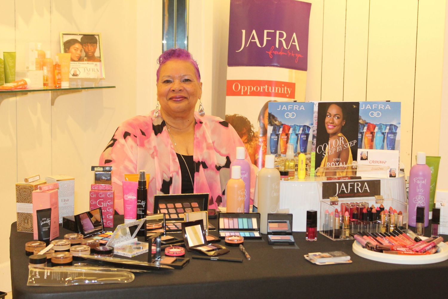Woman smiling behind a JAFRA cosmetics display table filled with makeup products at a promotional event.