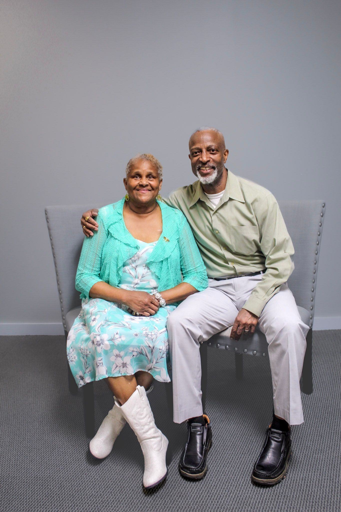 Smiling senior African American couple sitting together on a gray bench, wearing casual pastel clothing, posing for a portrait.