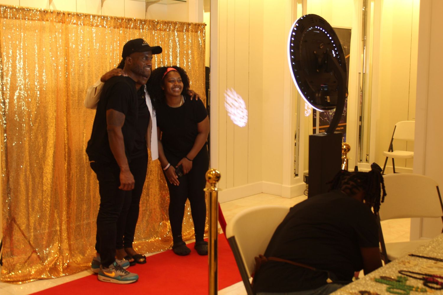 Two people posing and smiling in front of a gold sequin photo booth backdrop on a red carpet at an event.