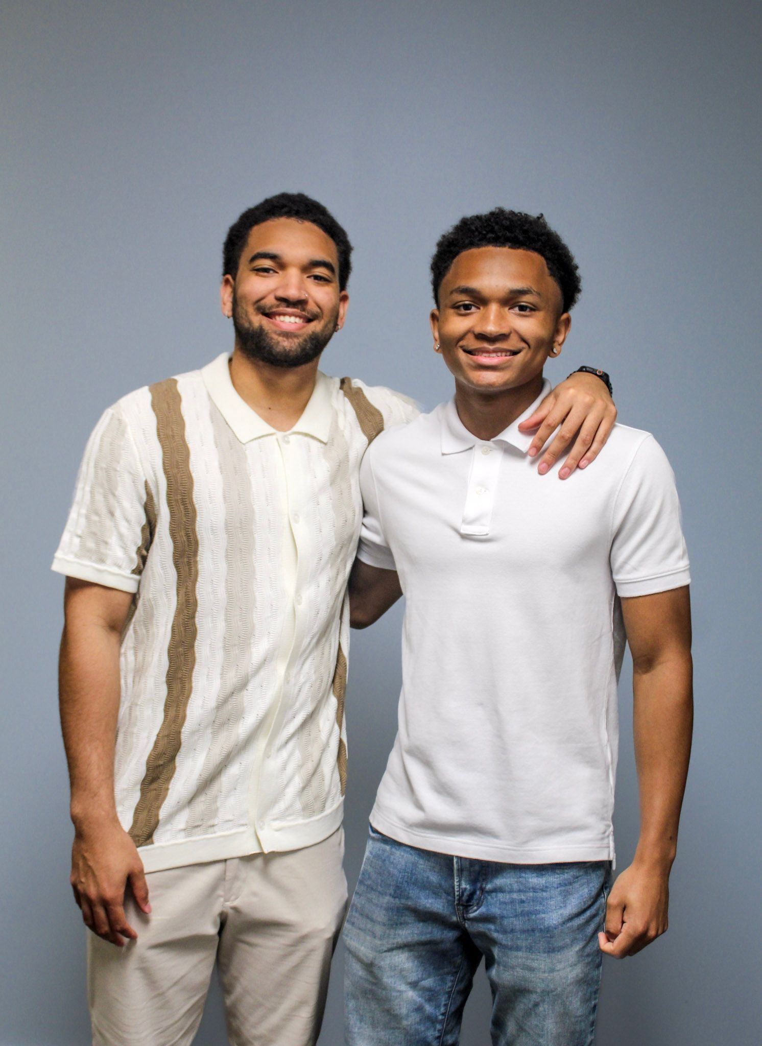 Two young men smiling and posing together, one in a striped polo and one in a white polo shirt, against a gray background.