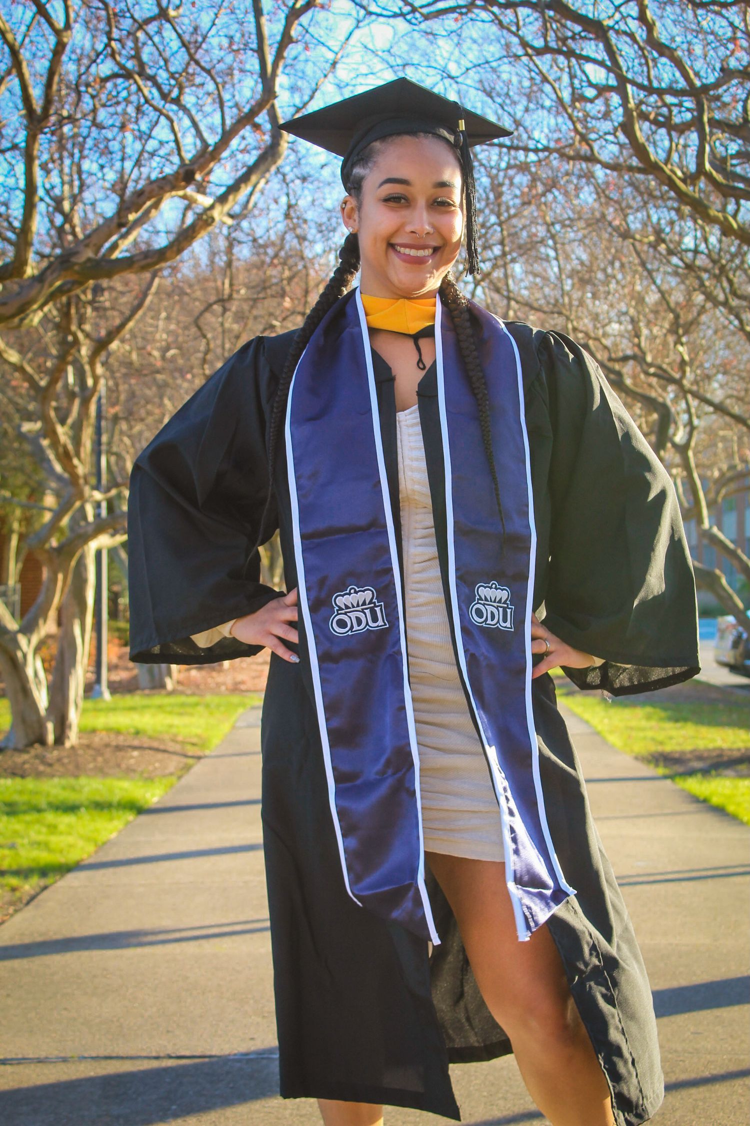 A smiling graduate poses in black cap and gown with ODU blue stole on a sunny campus walkway.