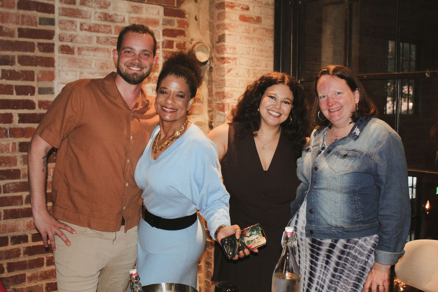 Four smiling people posing together at a casual indoor event near a brick wall.