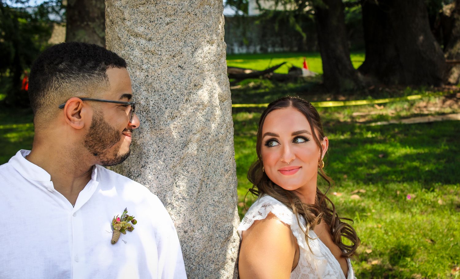 Bride and groom lean against a tree in a sunny park, smiling at each other on their wedding day.