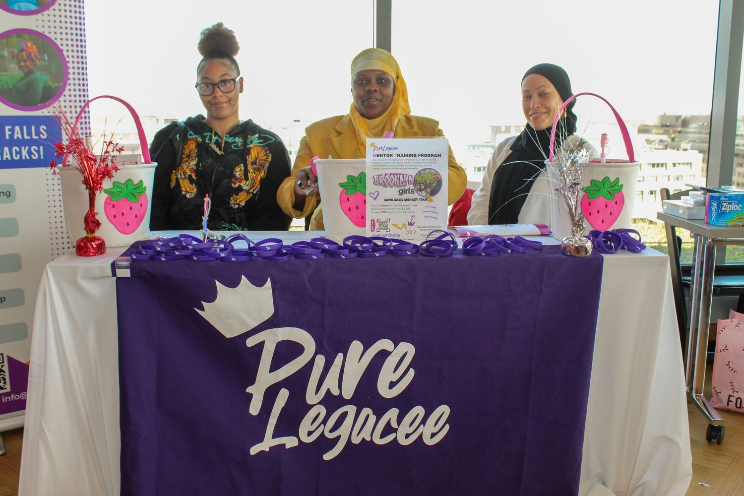 Three women stand behind a purple Pure Legacee branded table display with strawberry decorations at an indoor event.