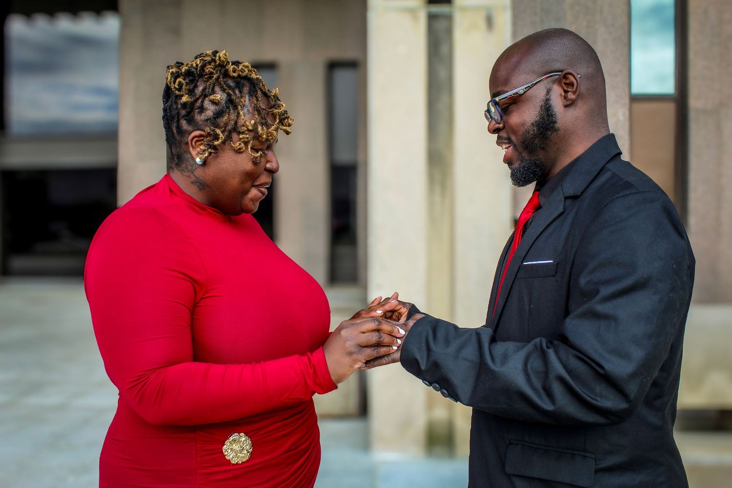 A man in a black suit places a ring on a woman in a red dress during an outdoor engagement ceremony.