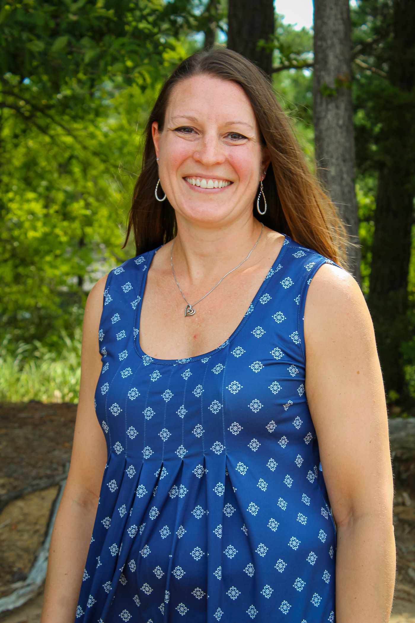 Person in blue patterned sleeveless top poses outdoors among green trees.