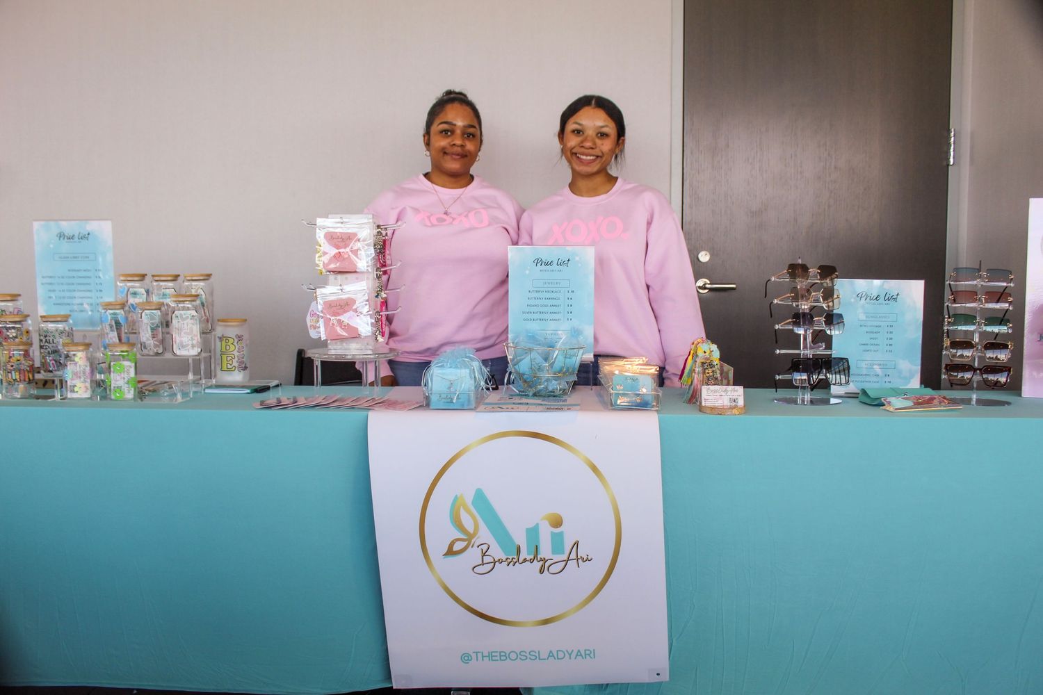 Two women in pink sweatshirts smile behind a teal vendor table displaying candy and sweets at a market event.