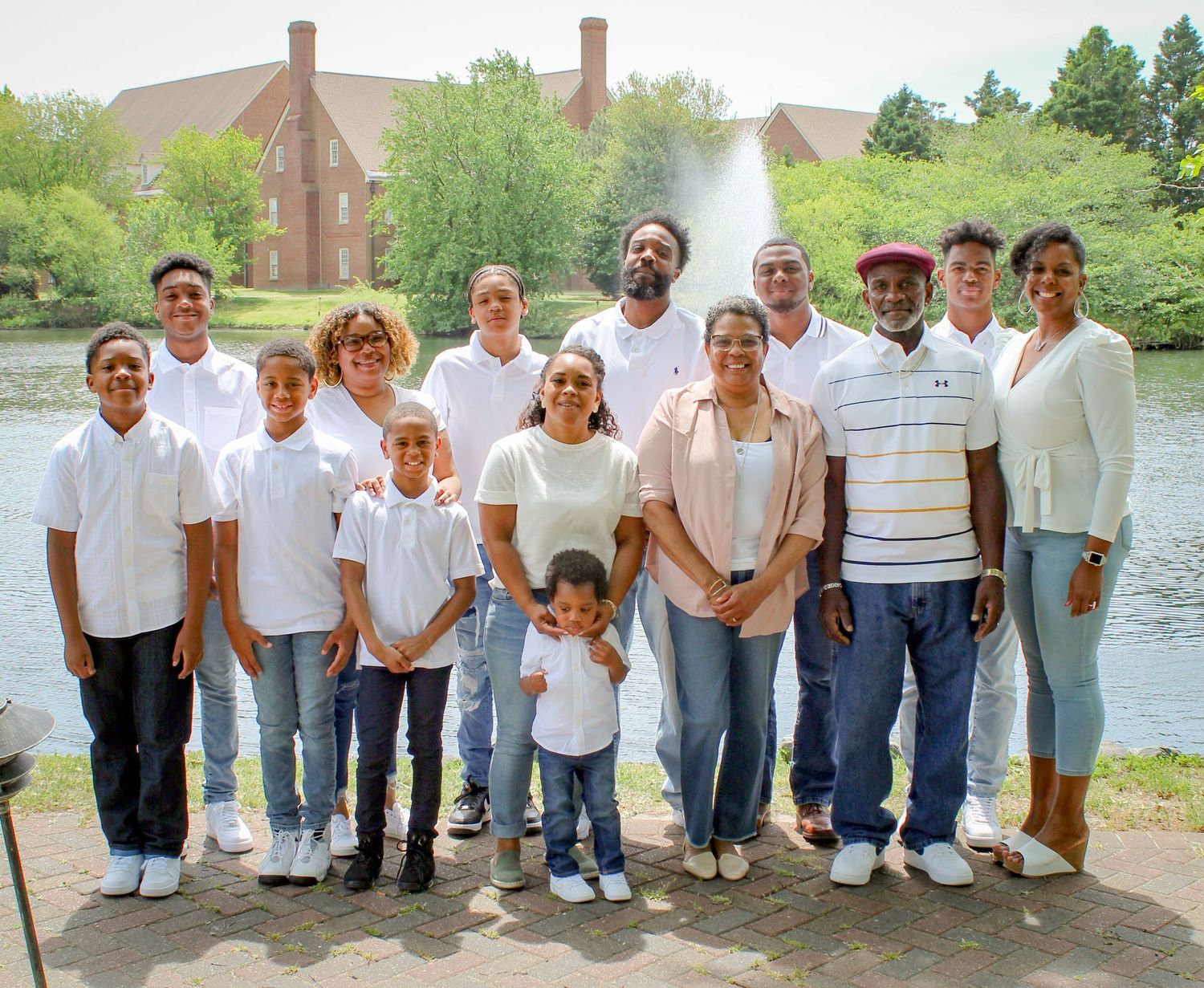 Large family group portrait outdoors near a pond with a fountain, dressed in white tops and jeans.