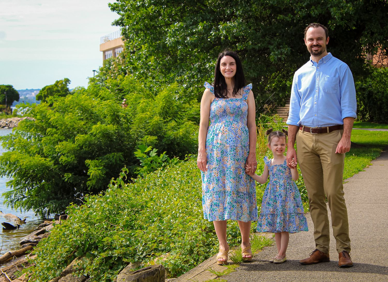 Pregnant woman in floral dress stands with husband and young daughter by waterfront park on sunny day.