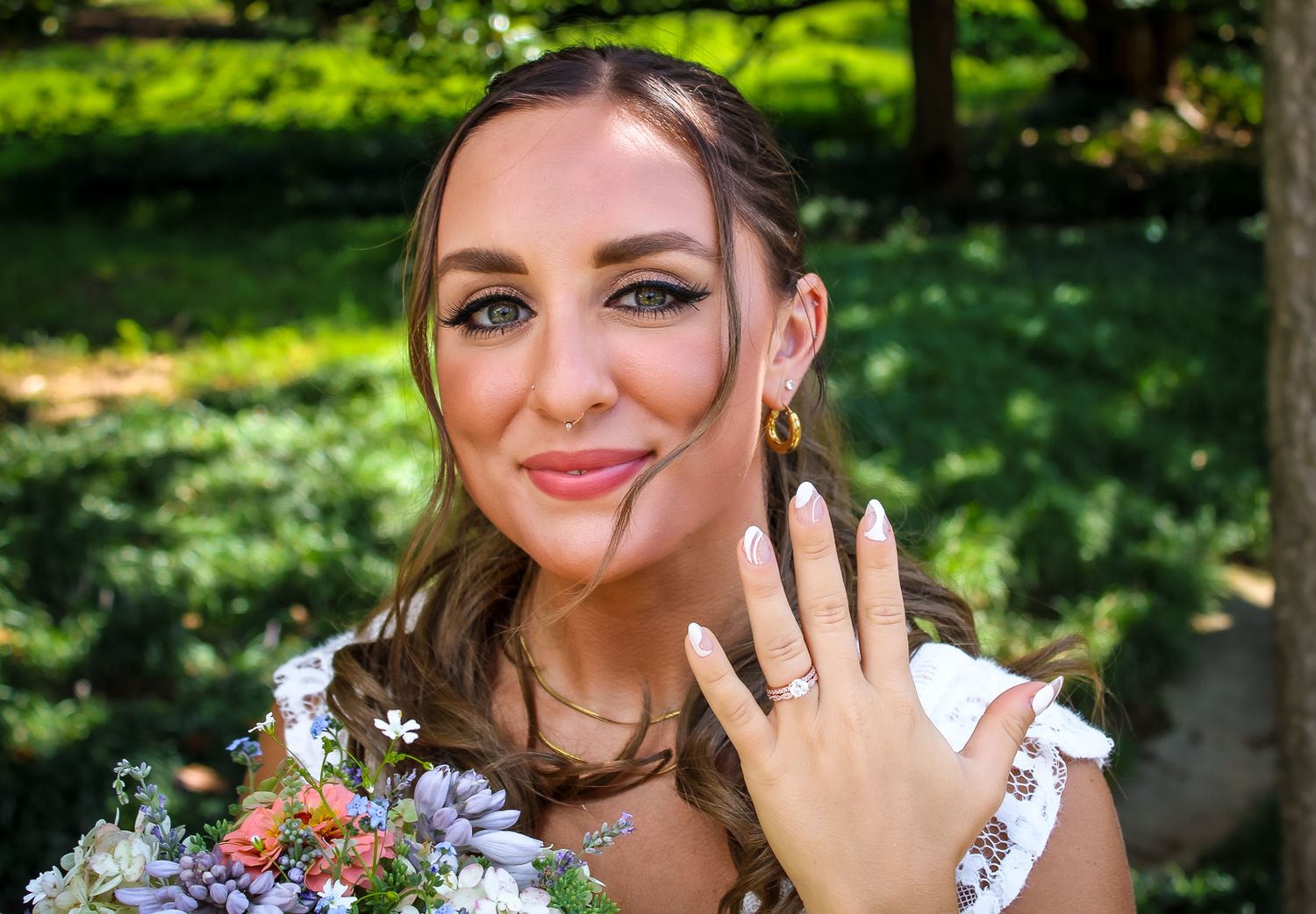 Smiling bride in white dress showing off engagement ring while holding a colorful bouquet outdoors.