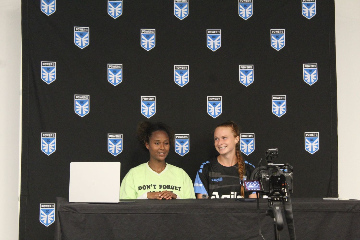Two women smile at a press conference table in front of a black backdrop with shield logos.