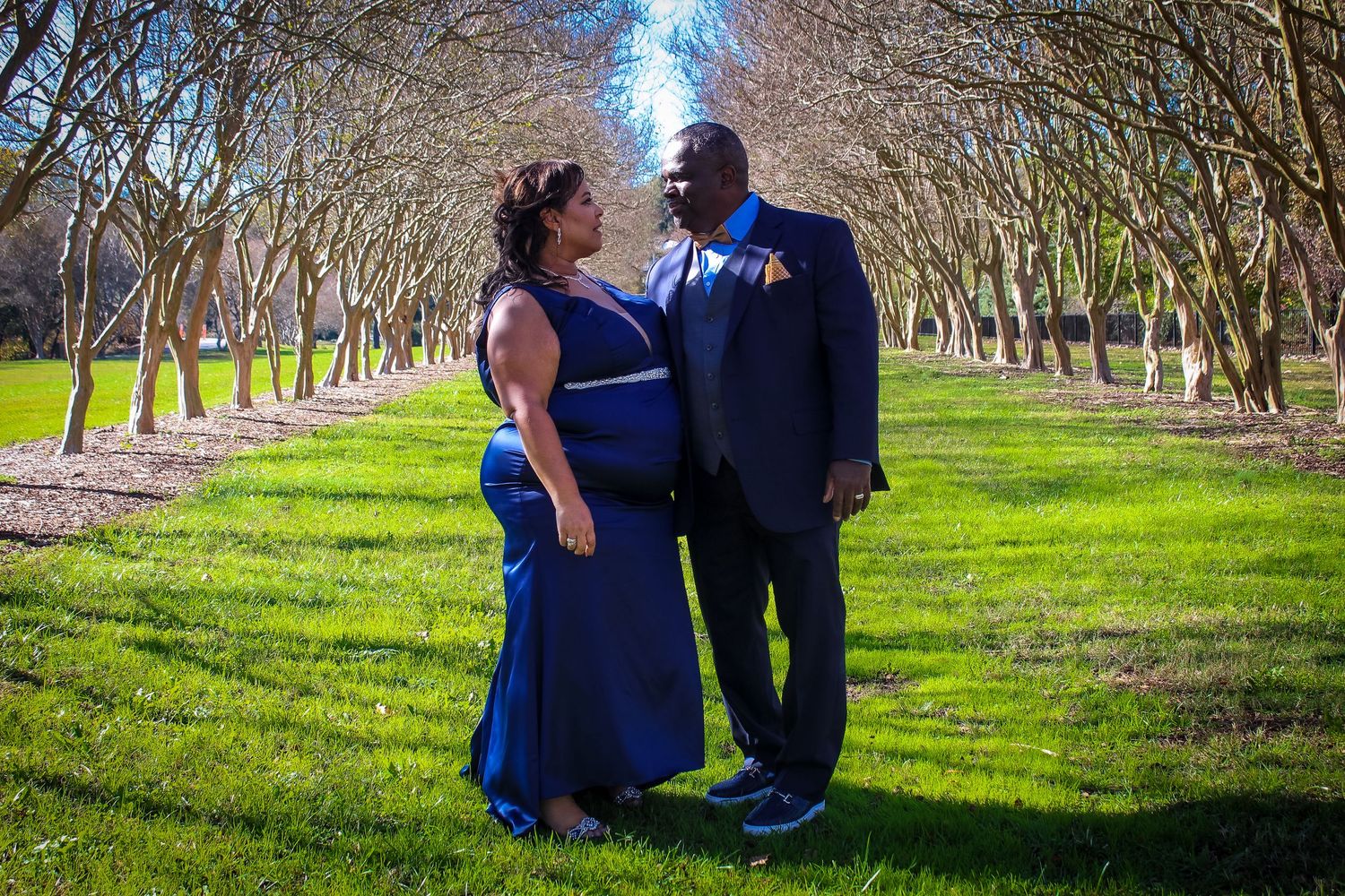 Couple in blue formal attire standing in a lush green park with bare trees in the background.