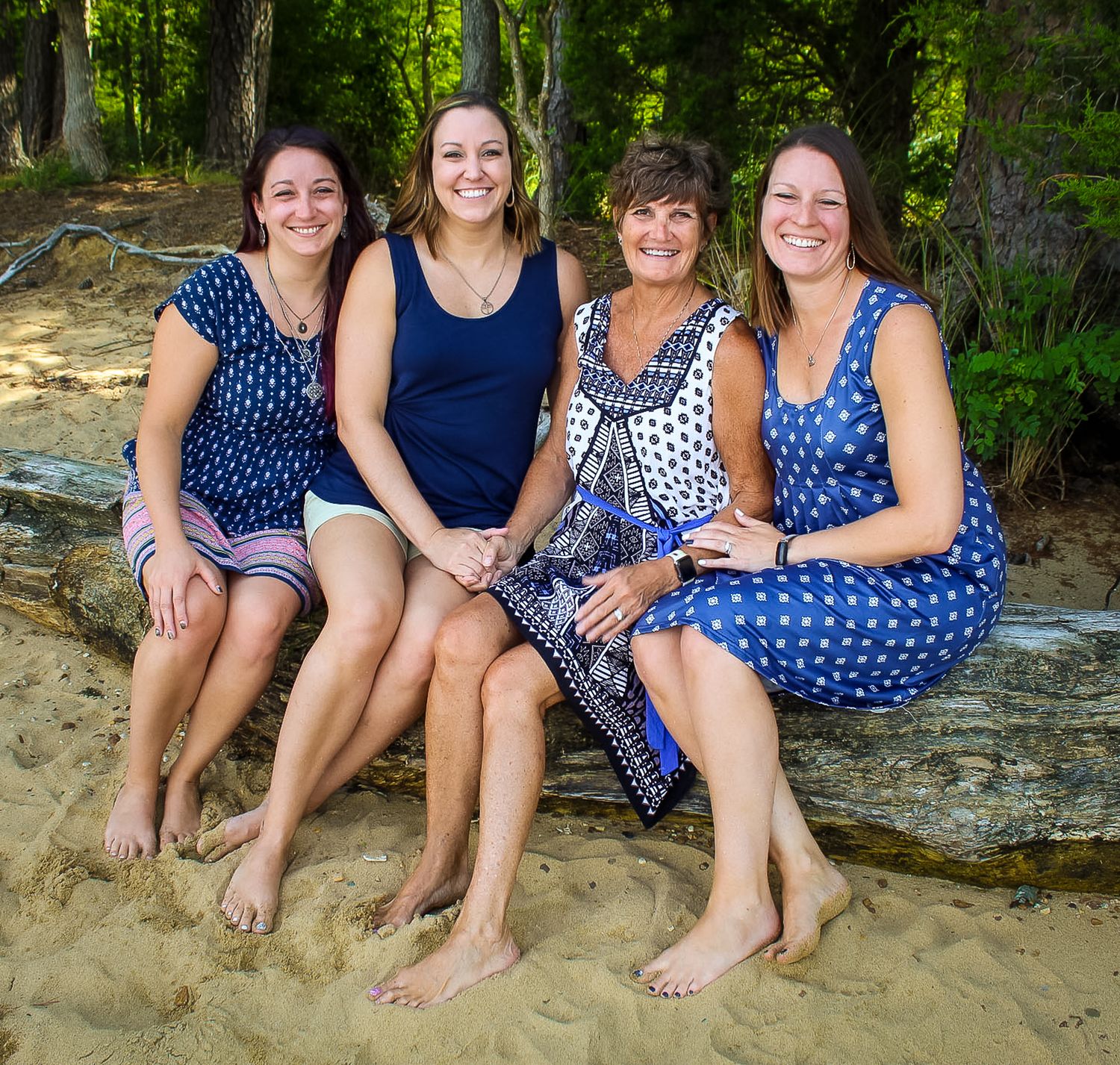 Four women sitting on a rock on a sandy beach, smiling, wearing blue outfits, with trees in the background.