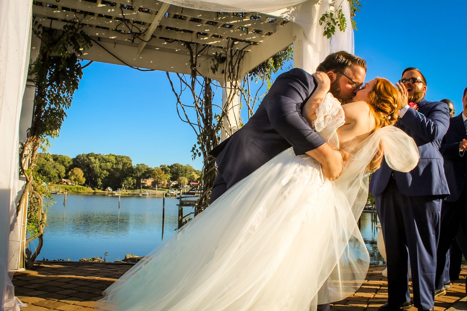 Bride and groom share first kiss at outdoor lakeside wedding ceremony under white floral arbor on sunny day.