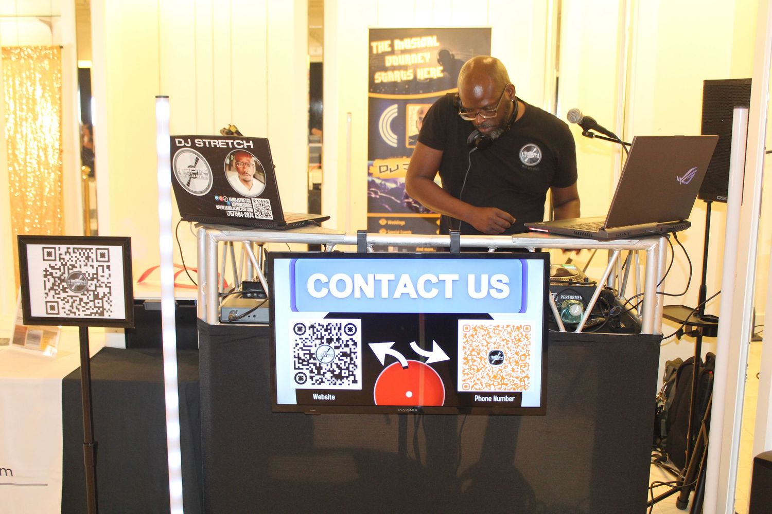 DJ Stinton at a professional DJ booth with a Contact Us sign at an indoor event expo.