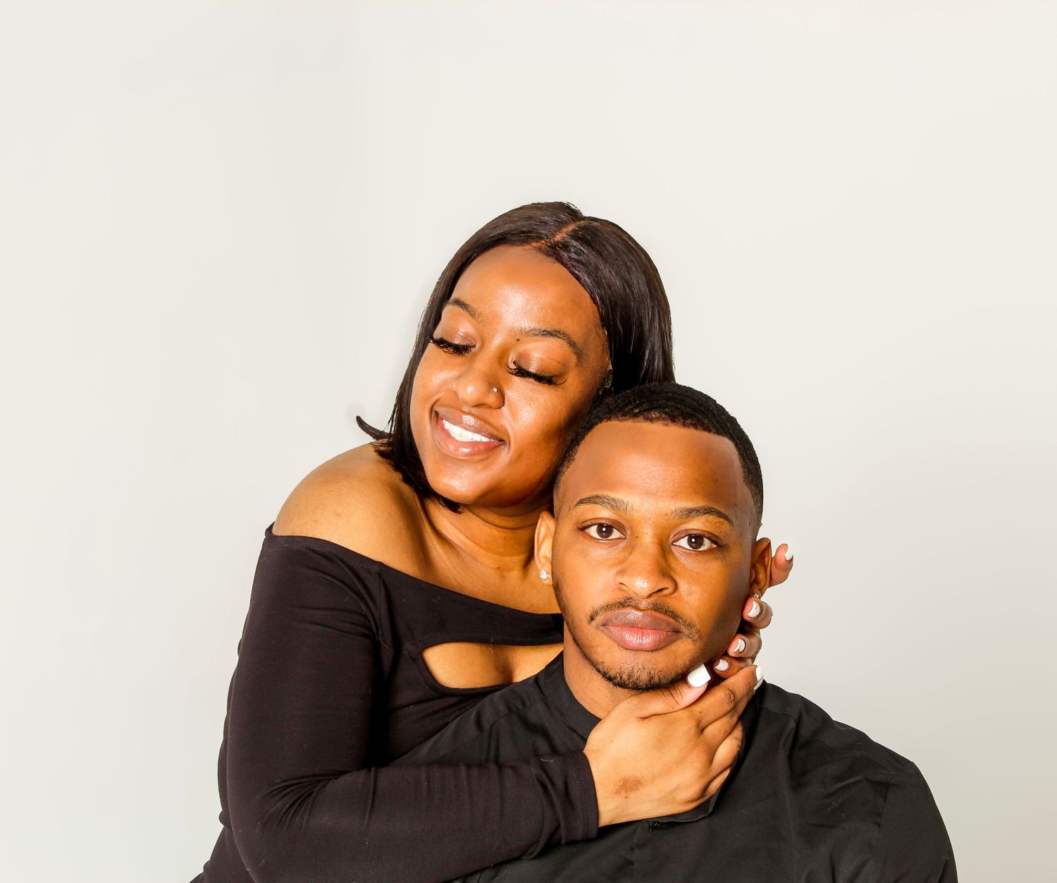 A smiling couple in black outfits poses together against a white background, woman embracing man from behind.