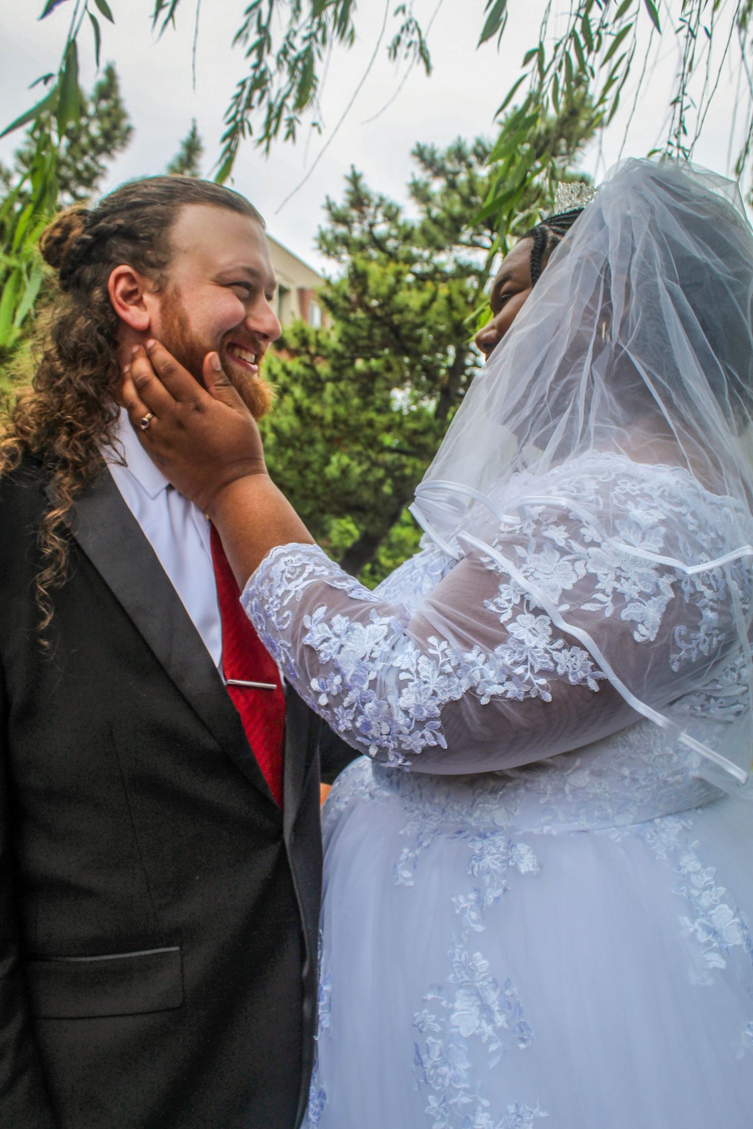 Bride in white lace gown and veil tenderly touches smiling groom's face outdoors at wedding ceremony.