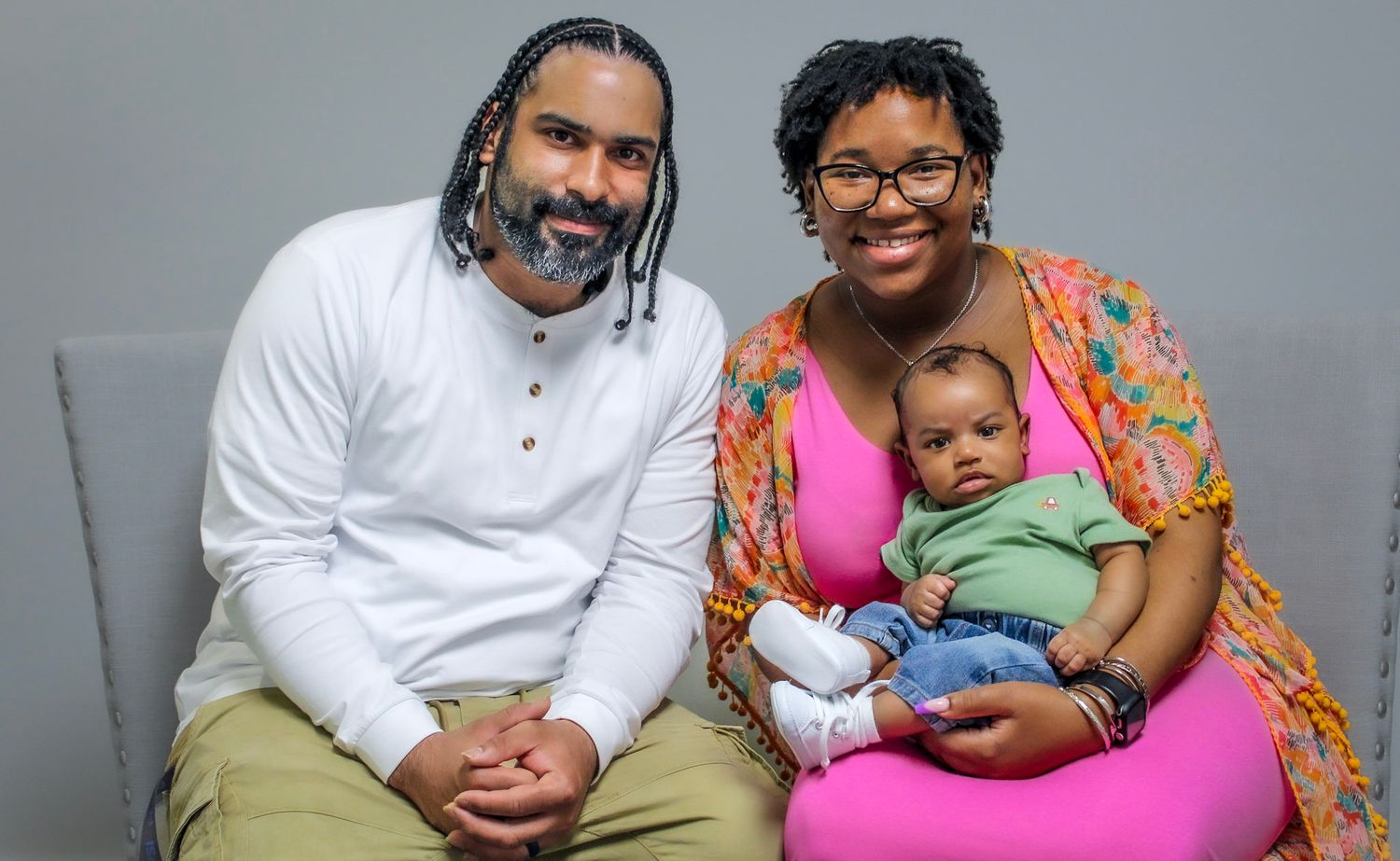A family portrait of two adults and a baby posing together against a gray studio background.