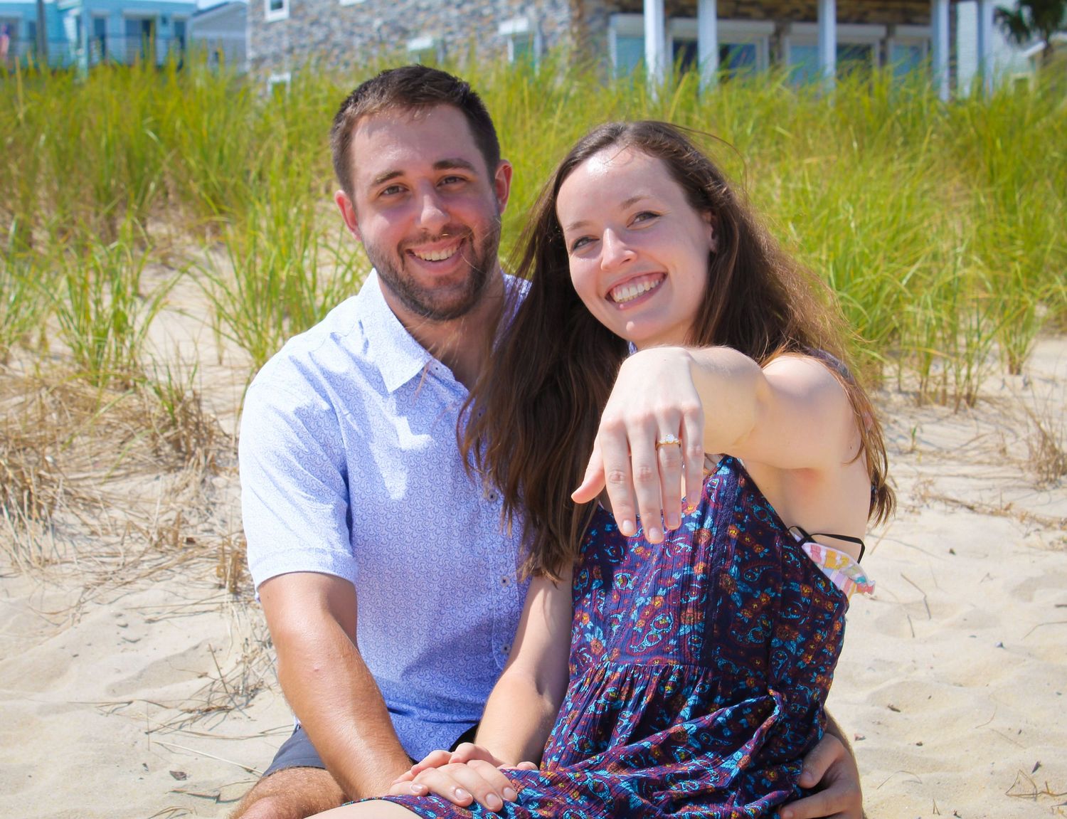 Happy couple sitting on sandy beach, woman showing engagement ring, smiling outdoors near coastal dunes.