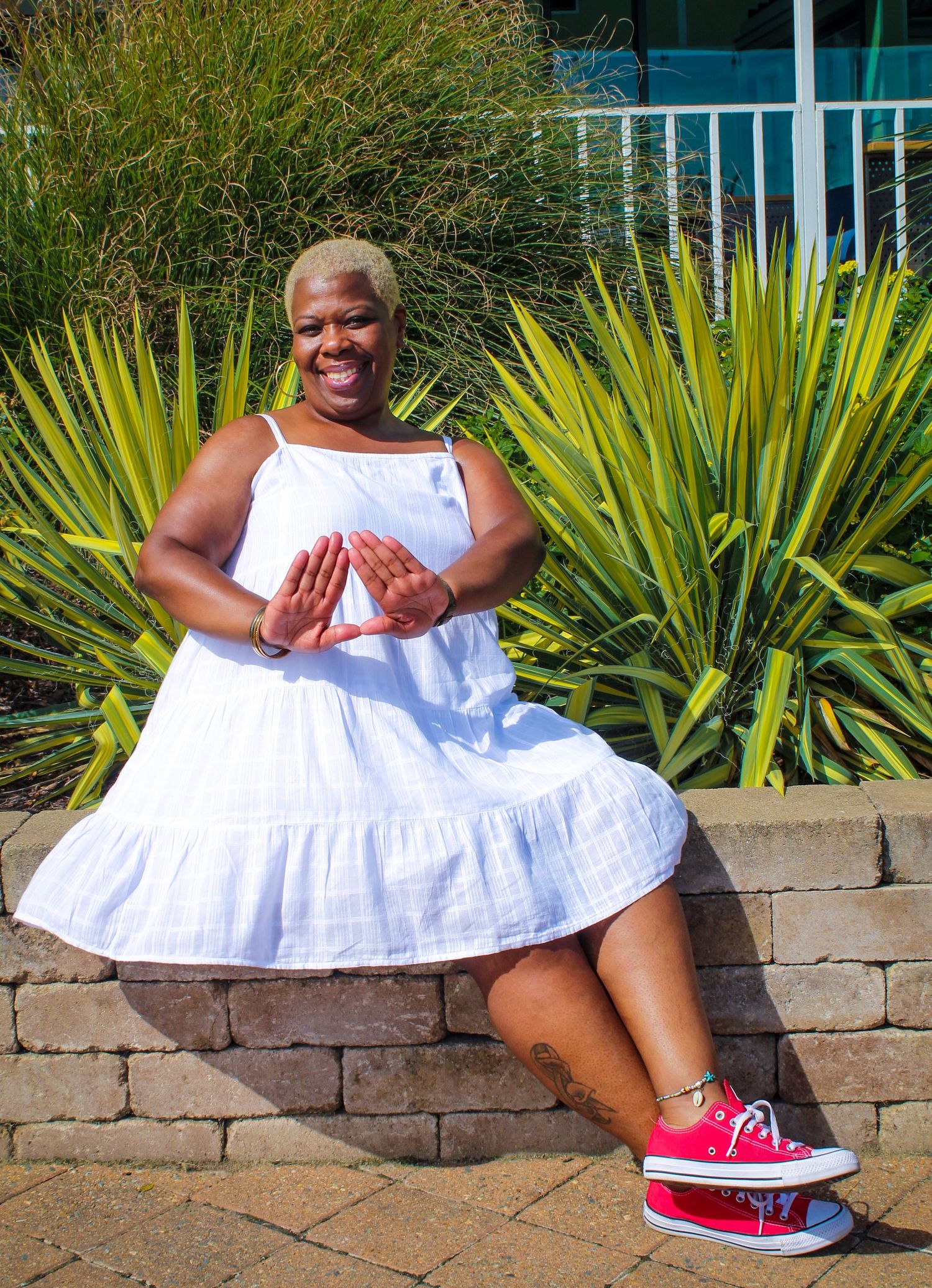 Person in white sundress and red sneakers sits on stone wall near tropical plants.