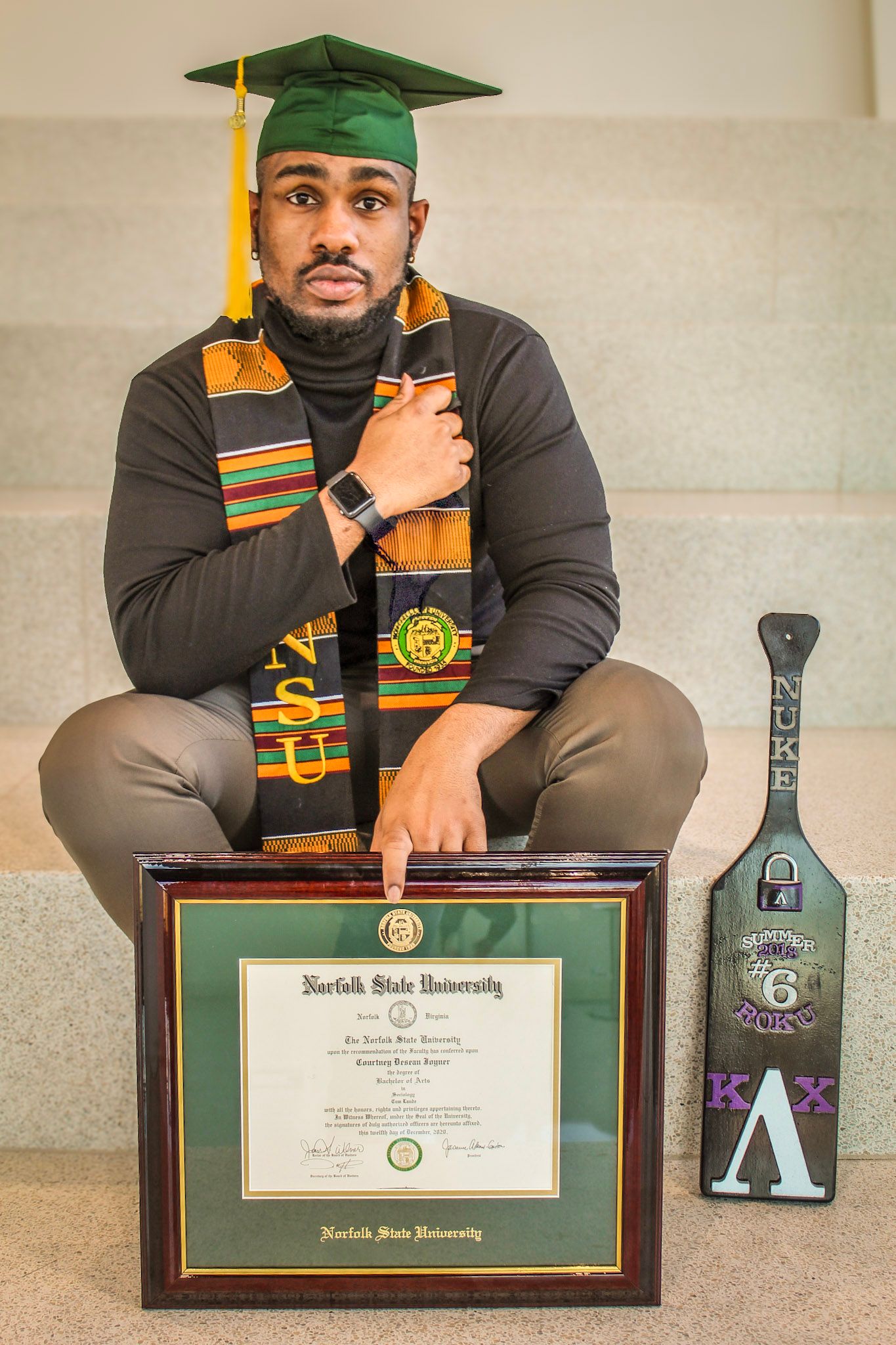 Graduate in green cap and gown poses with framed diploma and decorative stoles.