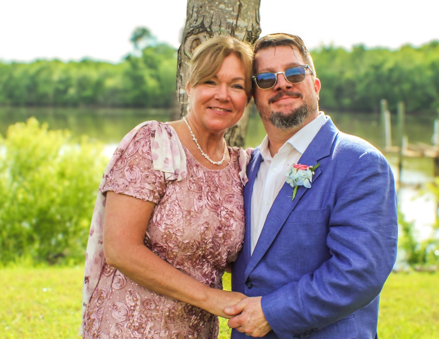 A couple in pink and blue formal attire embraces while celebrating outdoors near a waterfront on a sunny day.