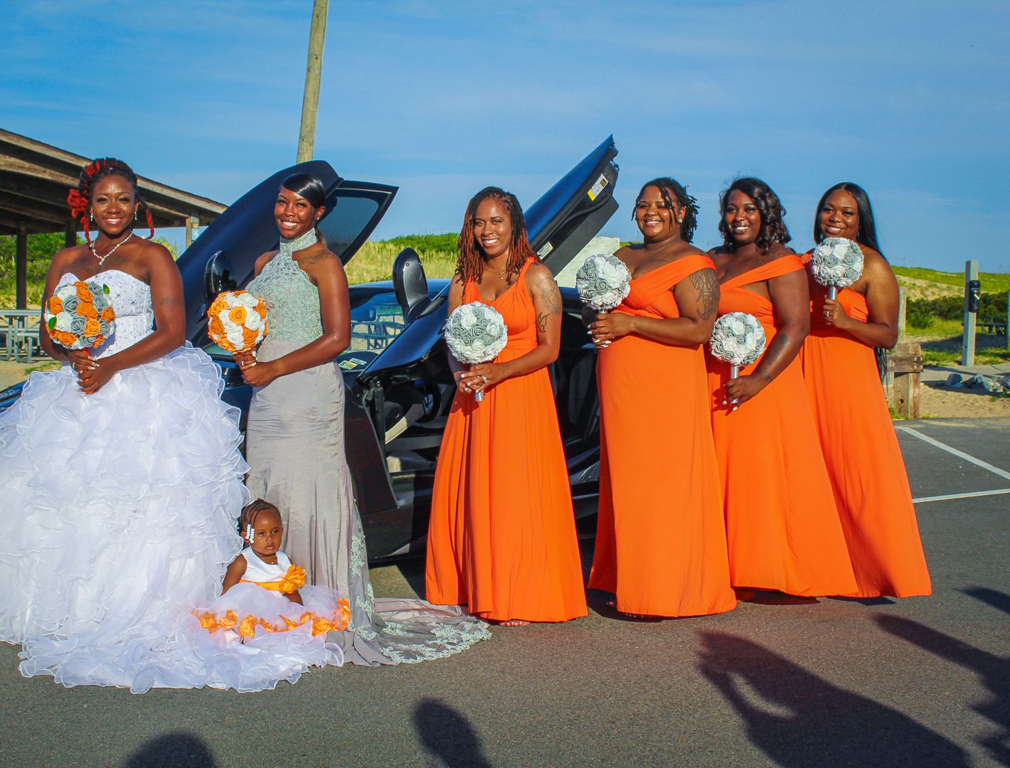 Bride in white gown with bridesmaids in orange dresses holding bouquets, posing beside a blue car outdoors.
