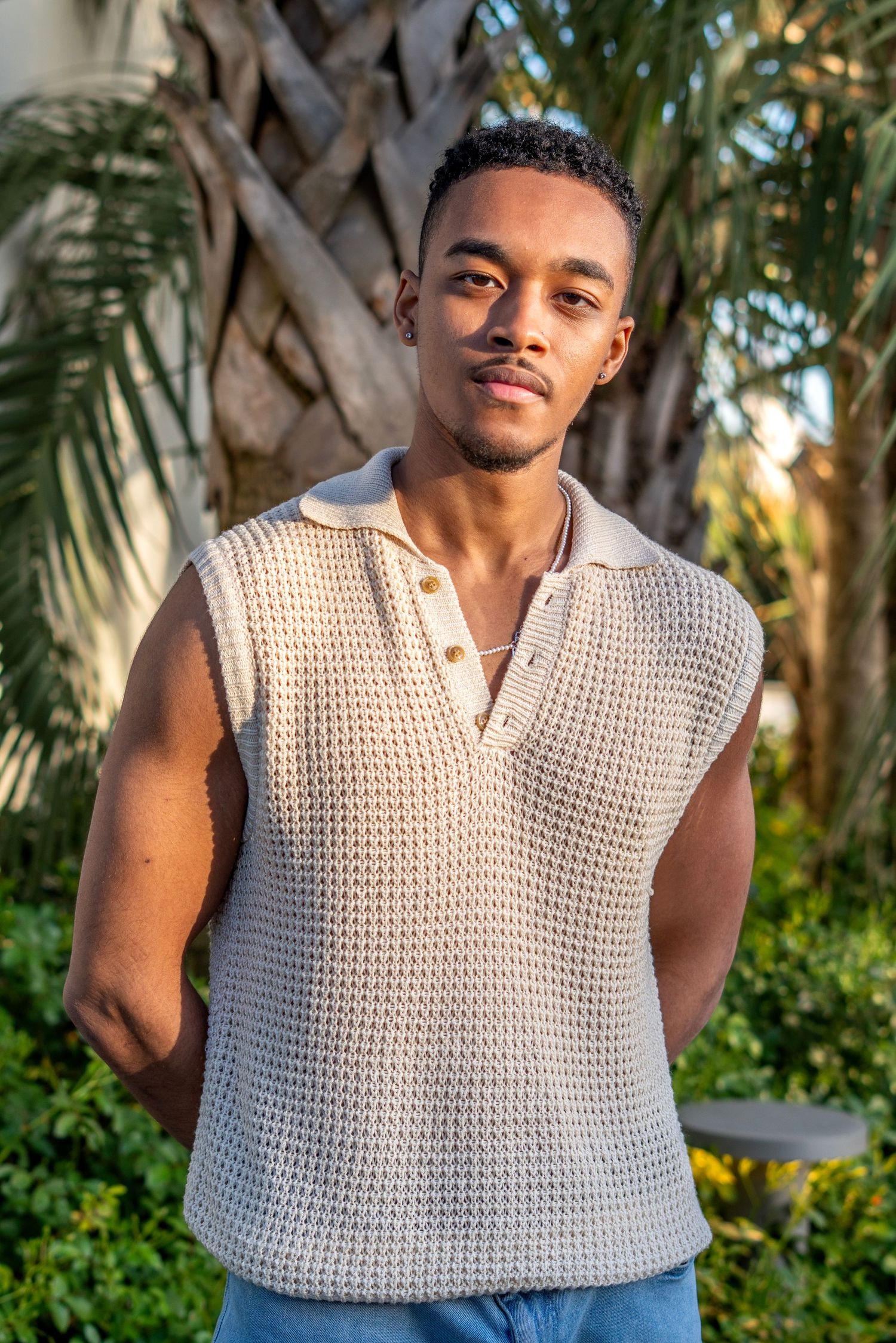 Young Black man in a beige crochet sleeveless polo shirt poses outdoors near tropical palm trees.