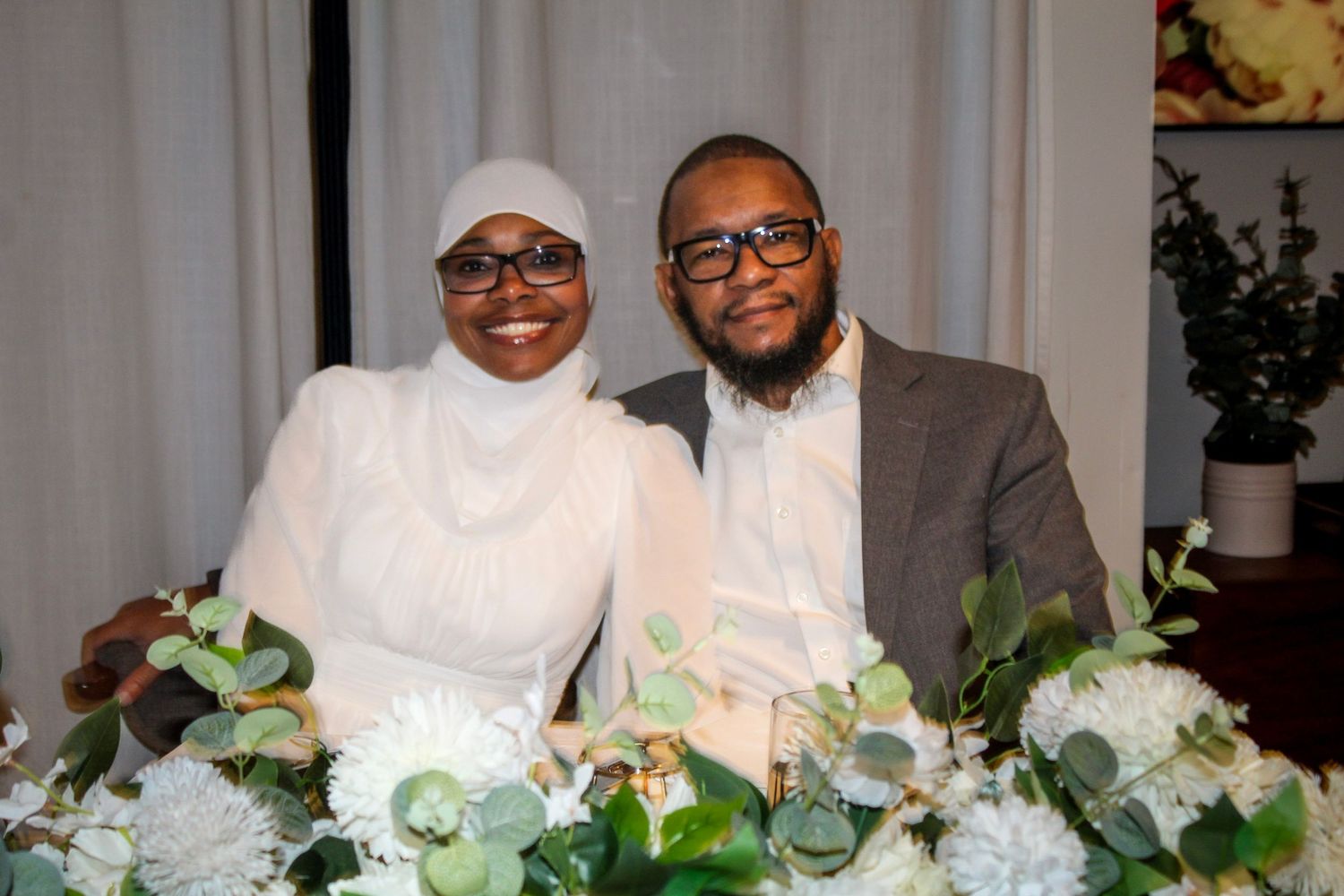 Couple smiling together at a floral-decorated table with white flowers and greenery at a formal event.