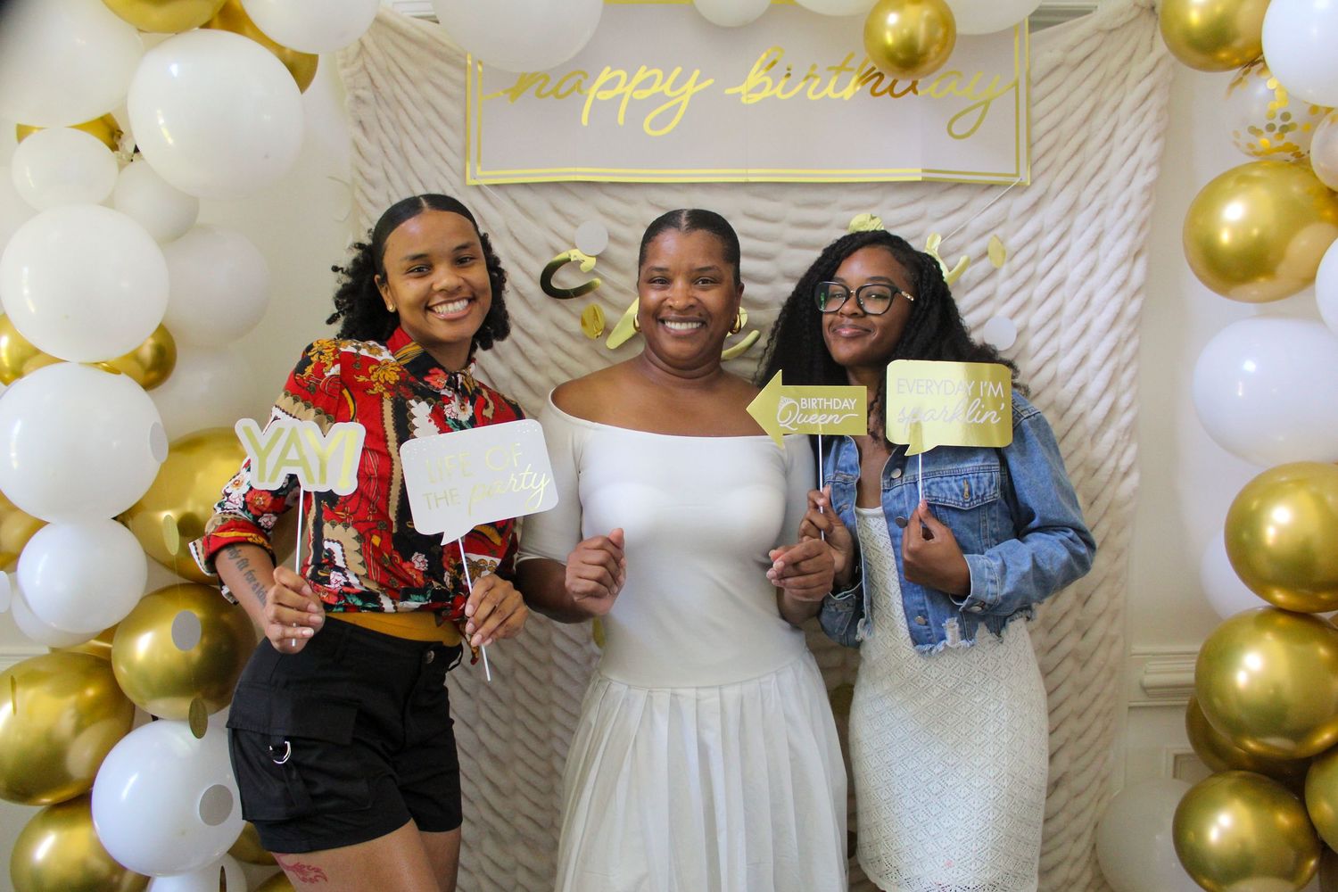 Three people pose with a festive balloon arch backdrop at a celebration.