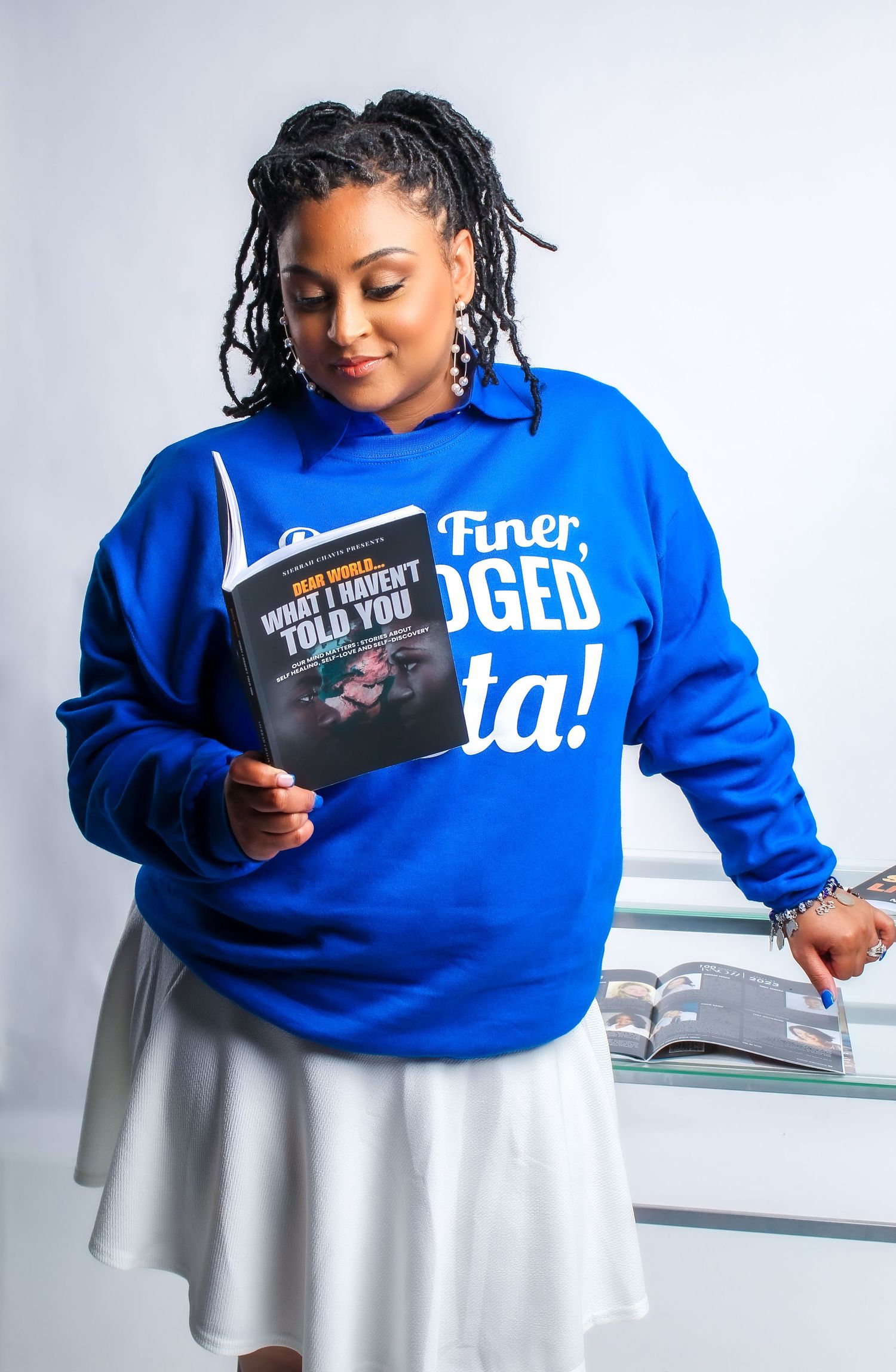 Woman in blue sweatshirt holding a book titled What I Haven't Told You, wearing a white skirt.