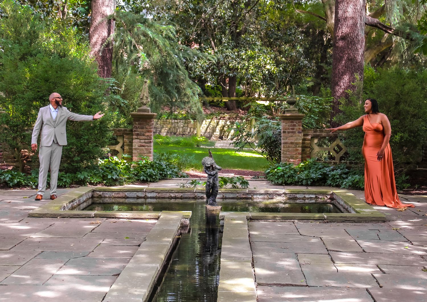 Two people pose in an elegant garden with a reflecting pool, brick walls, and lush green trees.