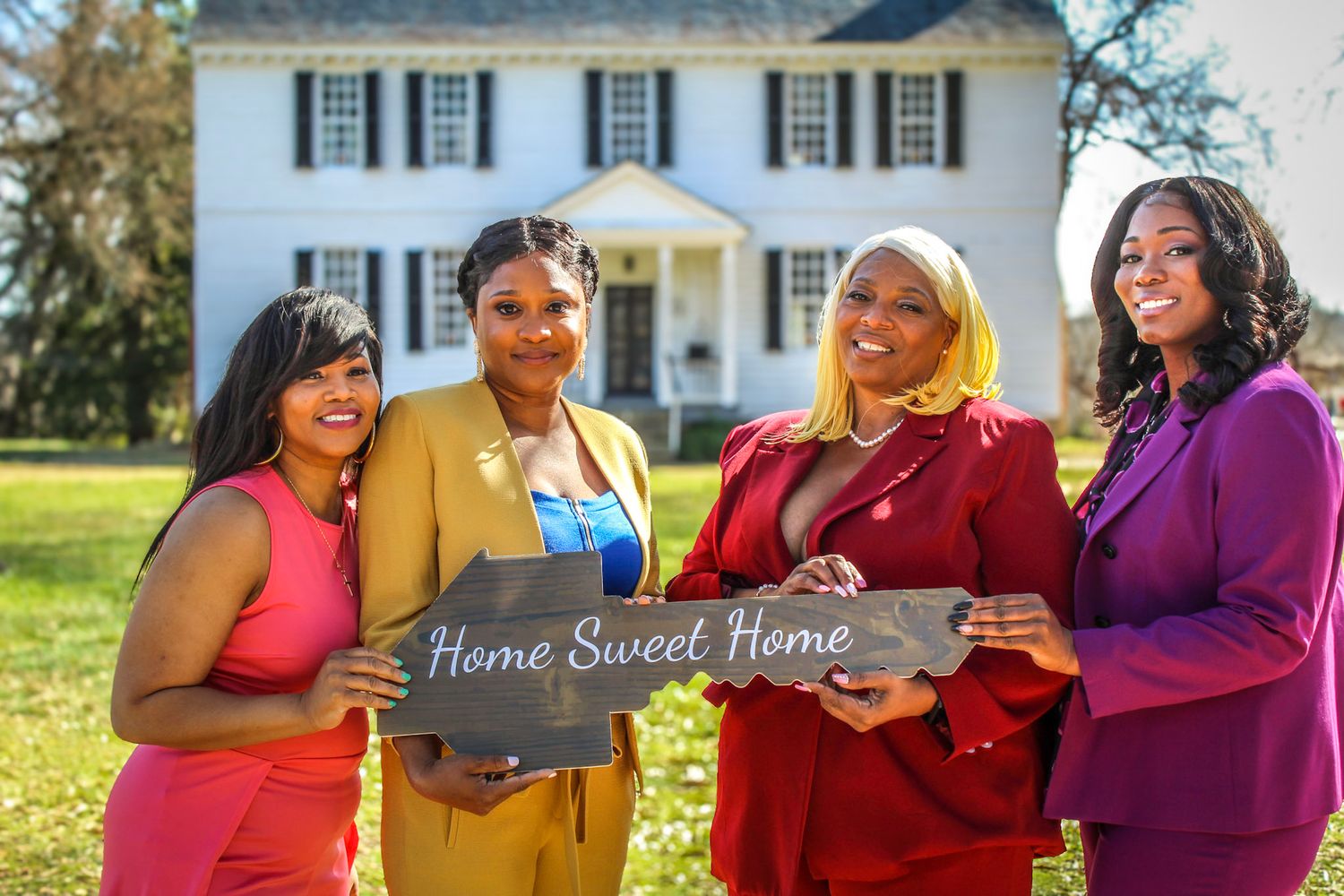 Group of friends in colorful outfits gather around a table outdoors with a white house in the background.