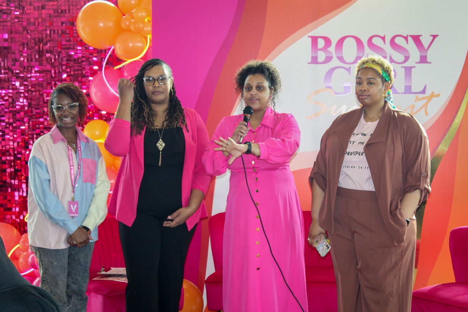 Four women stand on stage at a Bossy Girl event, with one speaking into a microphone against a pink backdrop.