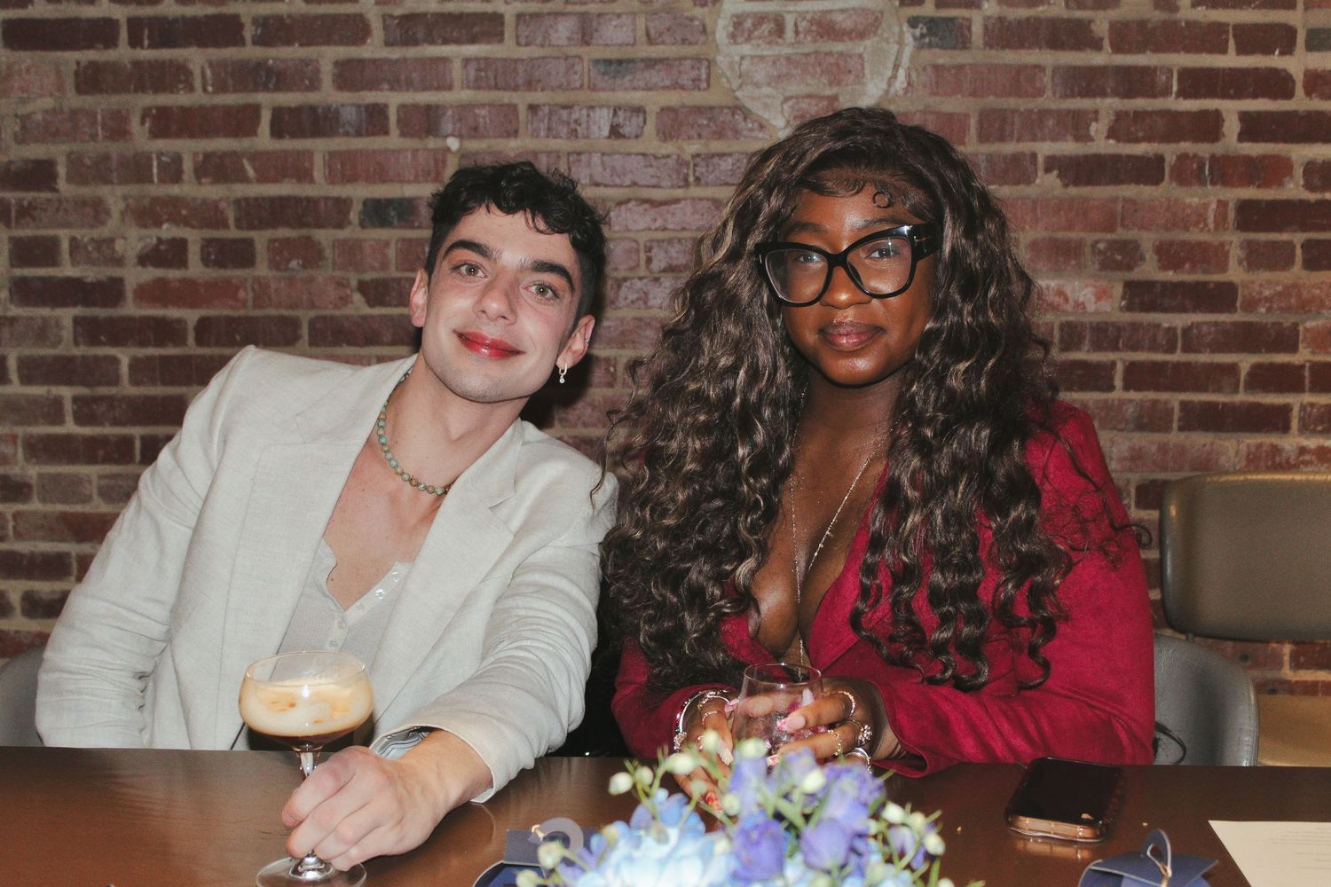 Two friends smiling together at a restaurant table with drinks and floral decor against a brick wall.