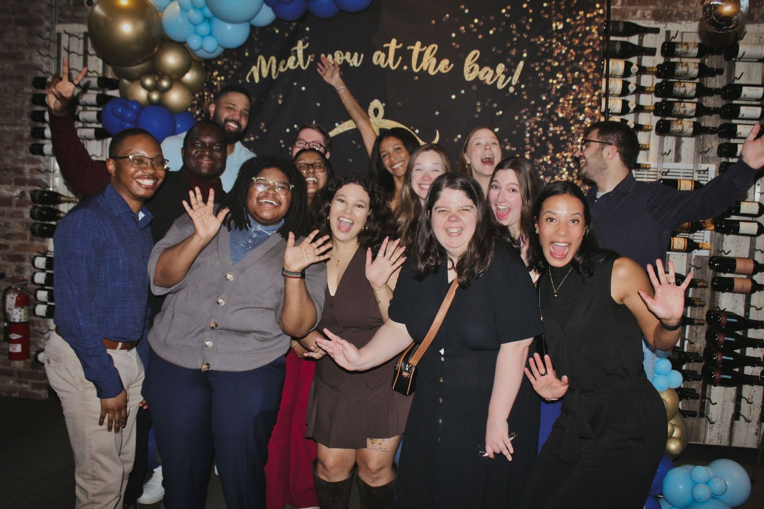 Group of friends posing and smiling at a bar event with balloon decorations and a Meet You at the Bar sign.