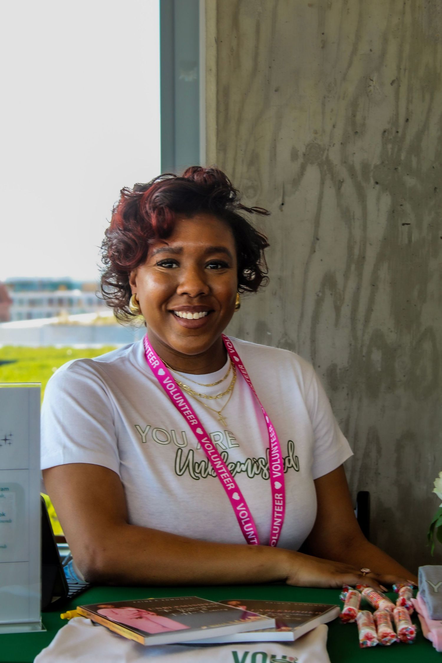 Smiling female volunteer in white t-shirt with pink lanyard seated at registration table at outdoor event.